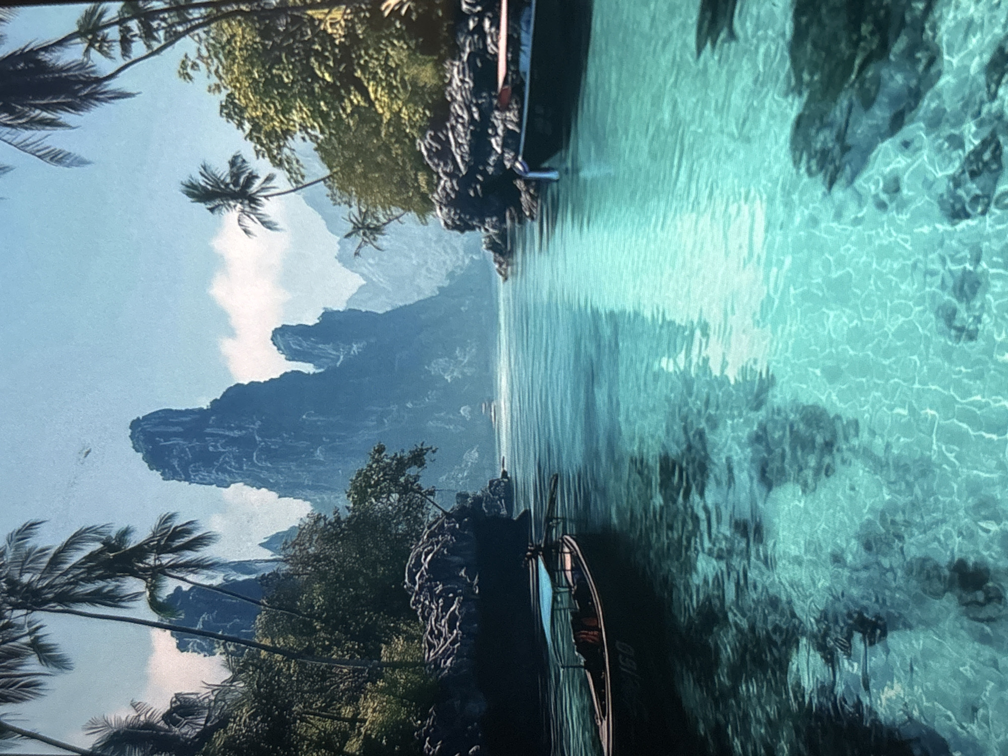 The image depicts a tropical scene with a narrow body of water, likely a river or inlet, flanked by lush vegetation and rocky cliffs. Tall, jagged mountains are visible in the background under a partly cloudy sky. Palm trees line the edges of the scene. Two boats are visible, one near the top and another near the bottom of the image, along the dark rock formations. The water is clear and turquoise, with visible patterns of sunlight reflecting on the sandy bottom. The location is not in Belopoptsi, Bulgaria.