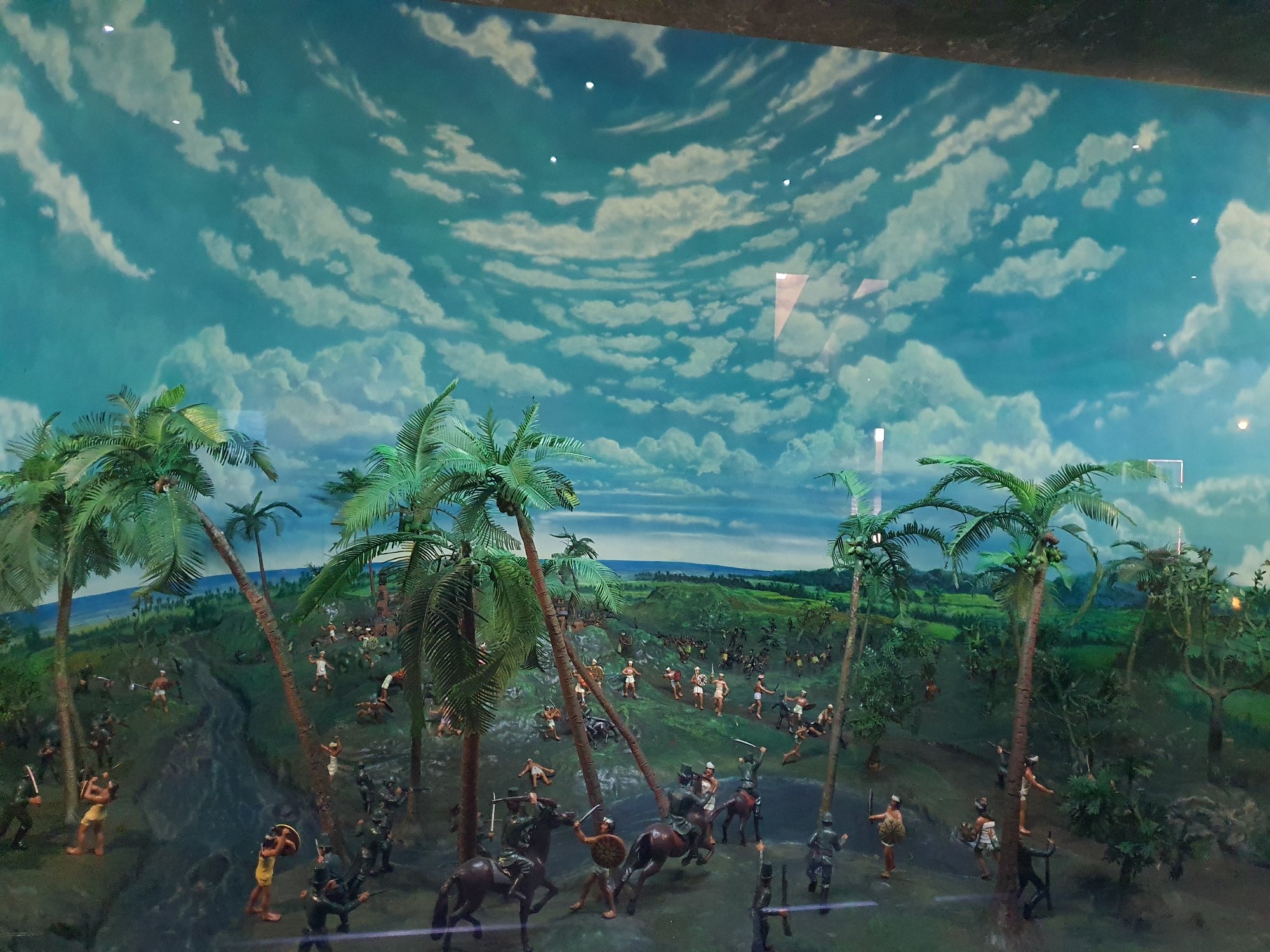 This is a diorama depicting a historical battle scene set in a tropical environment, likely intended for display in a museum. The scene features numerous miniature figures representing soldiers engaged in combat. Palm trees are prominently displayed throughout the diorama, suggesting a Southeast Asian setting. The background is a painted sky with clouds.
The diorama is located in the National Museum of Indonesia in Central Jakarta, Indonesia.
