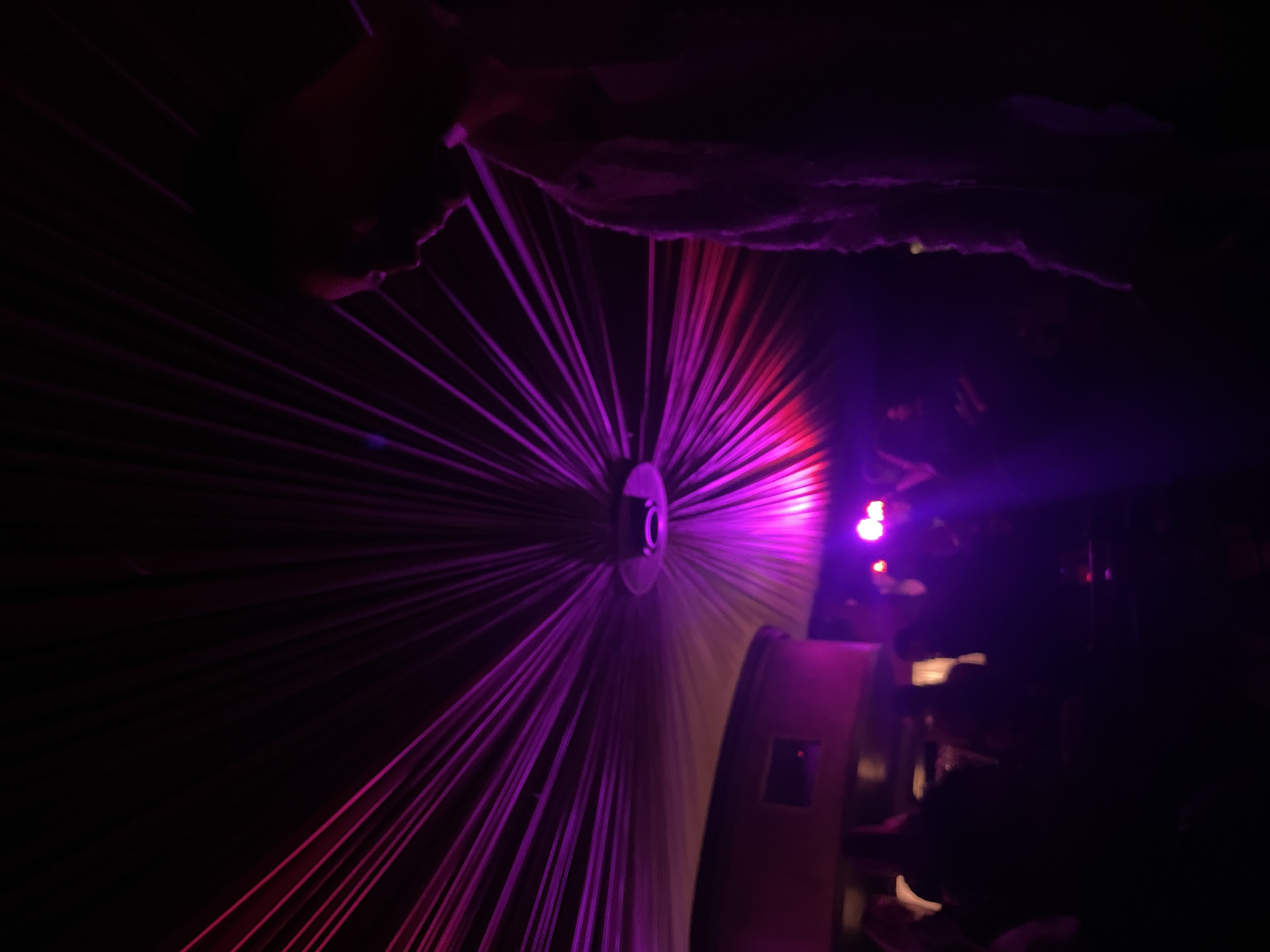 A concert or event takes place in a dark indoor space with a purple light show projecting from a central point with radiating lines. The equipment for the light show is visible along with some people in the background. The location cannot be determined from the image alone.