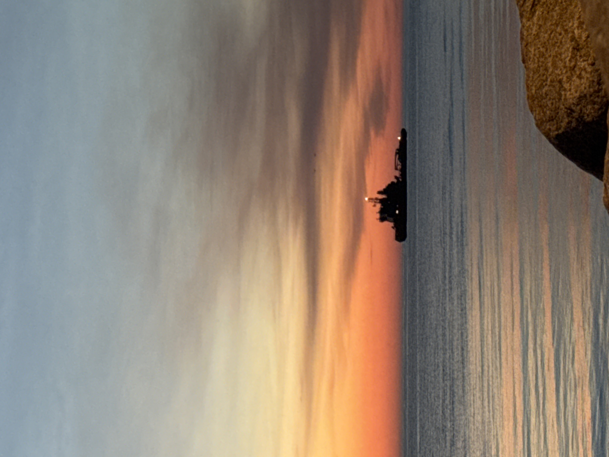 The image depicts a tranquil seascape at sunset with a cargo ship sailing on the horizon. The sky is painted in hues of blue, orange, and yellow, reflecting softly on the calm water. The ship, silhouetted against the vibrant sky, is positioned in the middle ground. In the foreground, a large rock is visible on the right, suggesting a coastal setting. The overall atmosphere is serene and peaceful, capturing the beauty of nature during twilight. The location of the image is not specified in the city of Tallinn, Estonia, but it is a general seascape.