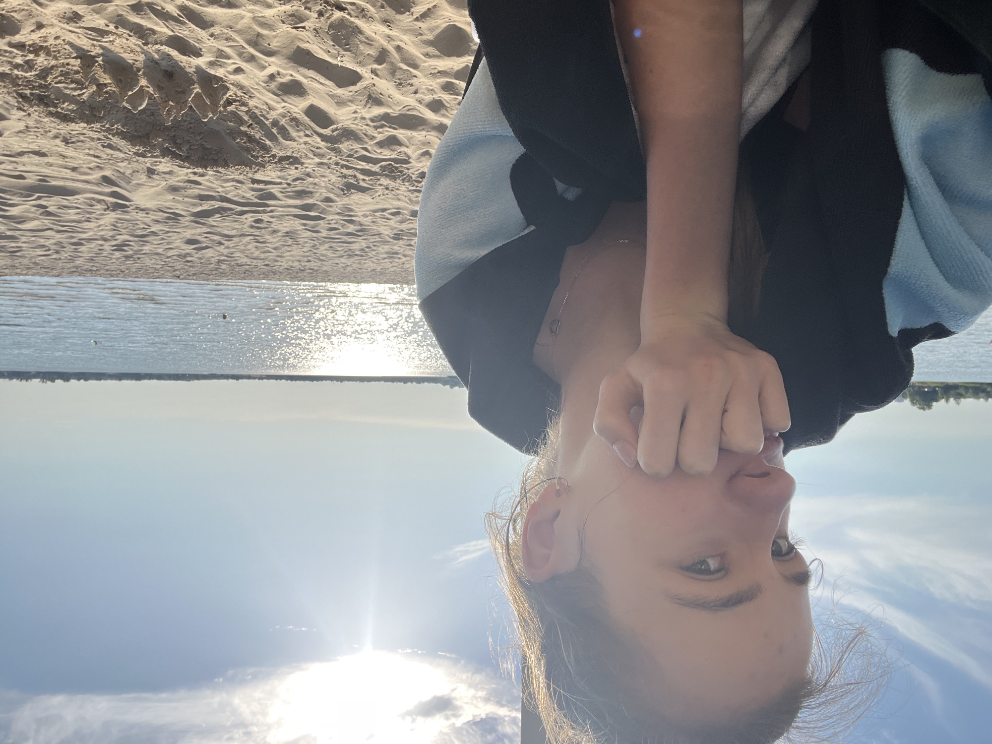 The media file depicts a young person with light brown hair and fair skin, captured in an upside-down orientation. They are wearing a black and light blue jacket. Their right hand is raised to their face, partially obscuring their mouth and nose, while their eyes are open and looking towards the viewer. A delicate silver necklace with a small charm is visible around their neck.
The background suggests an outdoor setting, likely a beach, with visible sand dunes in the upper portion of the image. Below the sand, a body of water, possibly a lake or sea, reflects the bright sunlight, creating a shimmering effect on its surface. A faint, distant tree line is visible on the horizon. The sky above is a bright blue with scattered white clouds. The strong sunlight and clear sky suggest it is daytime, likely late afternoon or early evening, given the warm tones in the water's reflection. The overall mood appears casual and relaxed. There is no visible text in the media file. The provided location context is Yelyzavetivka, Ukraine.