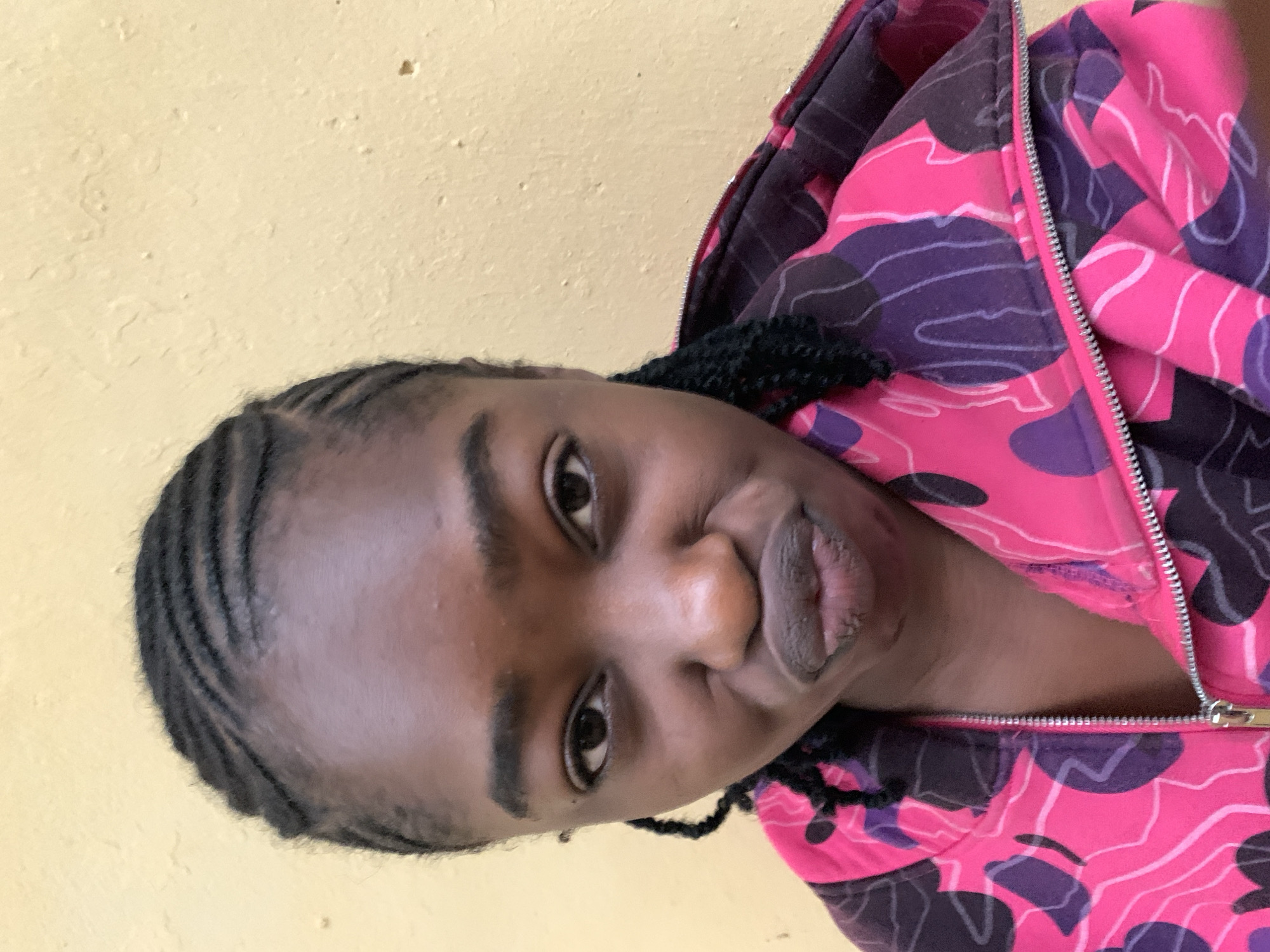 A close-up portrait photograph shows a young person, likely female, with dark skin and braided hair, looking directly at the camera. The image is oriented with the subject's head to the left and feet to the right. The person is wearing a vibrant pink and purple camouflage-patterned hoodie, featuring irregular shapes in varying shades of pink, purple, and black outlines, with a visible silver zipper running down the front. Their facial expression is a playful pout, with lips slightly puckered. The background is a textured, light yellow wall, suggesting an indoor setting. No specific location cues beyond the wall texture are visible within the frame. The lighting is soft and even, highlighting the details of the person's face and clothing. No text is visible in the image. The scene conveys a sense of casualness and playfulness. The location context is Rafin Par, Nigeria.