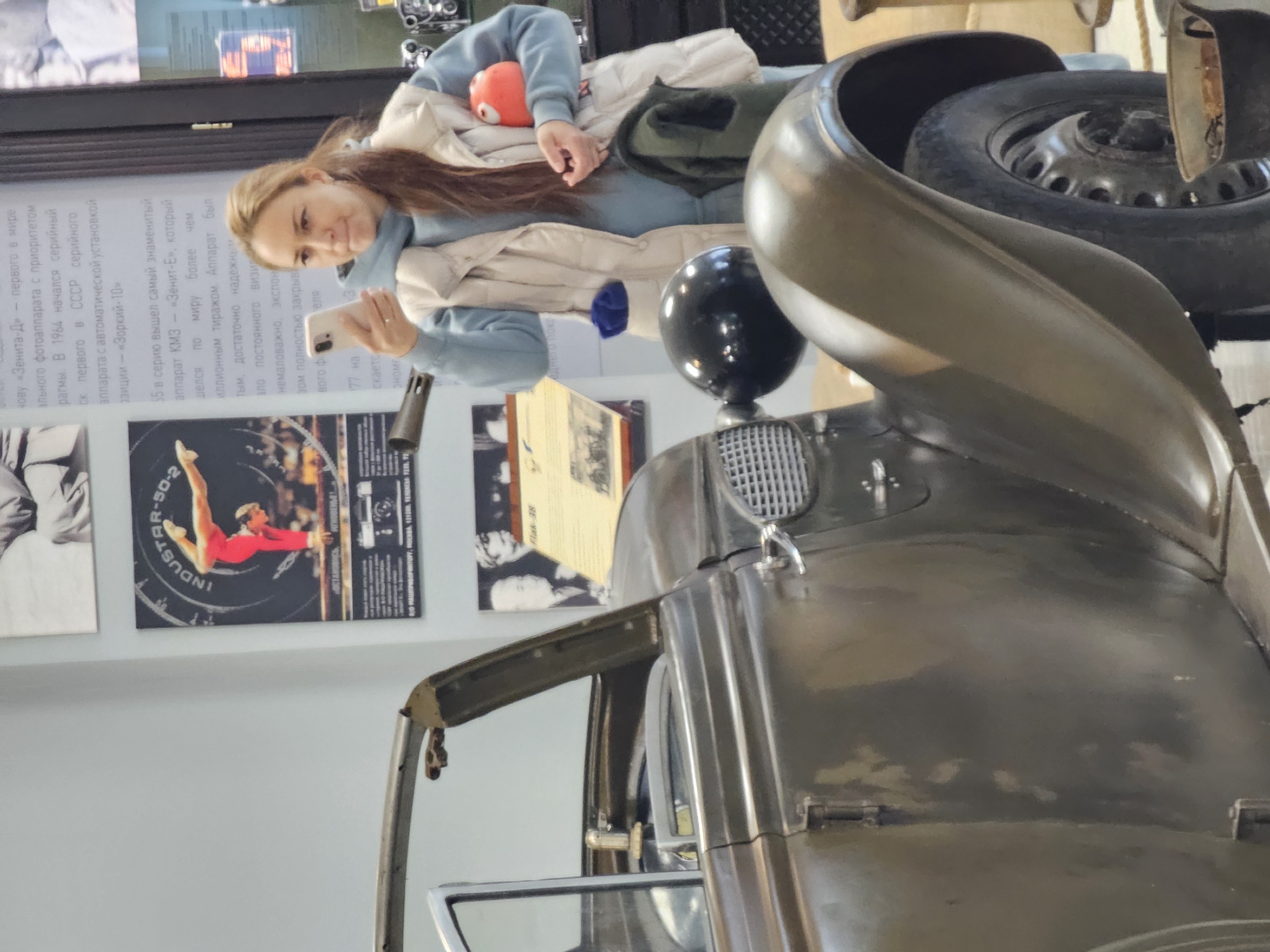 Here is a concise description of the image:
The image shows a woman in a light blue hoodie and beige jacket looking at her phone. She is standing near an old, olive-green vehicle, which appears to be a car. Behind her, there are informational posters on a wall, one of which features a photo of a gymnast. The OCR text suggests the location is in Russia, specifically referencing Moscow and the "Zenit-E" camera. Without additional context, it's impossible to confirm if the location is in the city of Архангельское, Russia.