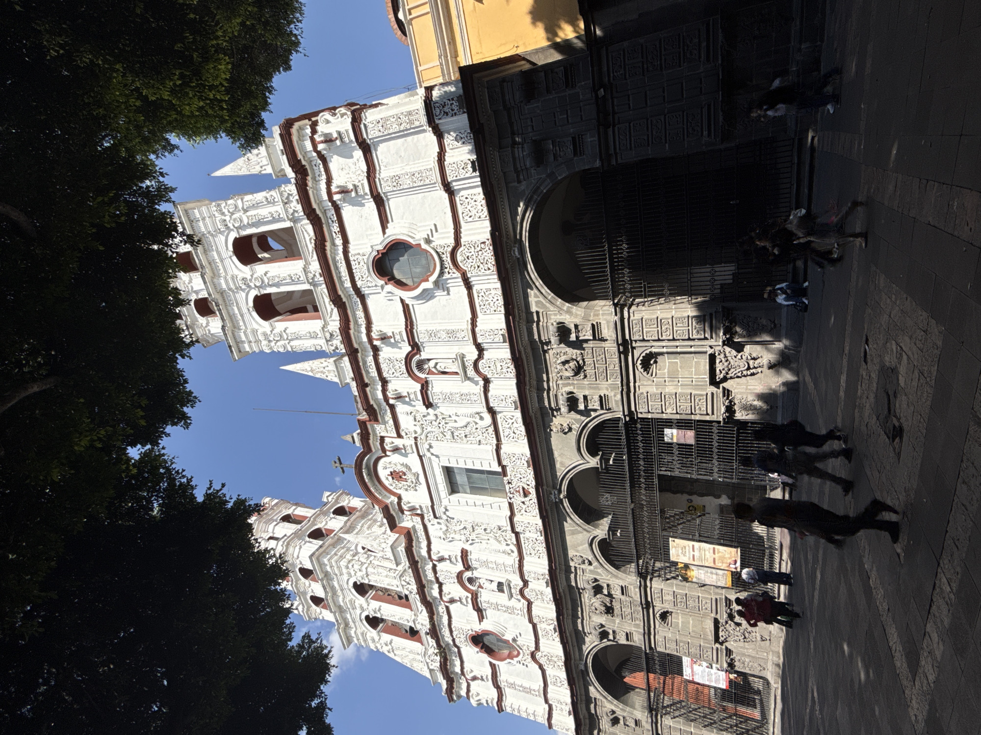 The image captures a street scene in Puebla, Mexico, featuring a distinct white building with ornate red detailing. The building is likely a church or cathedral due to its architectural style and the presence of a cross atop one of its towers. The building is white with complex decorative moldings, red trim around windows and along edges, and ornate details throughout the facade. The structure has multiple levels and features arched windows and doorways. The building is constructed with grey stone, with arches and ornate decorations. The entrance is covered with a dark metal gate. There are multiple flyers or signs posted on the entrance.
The building casts a long shadow onto the street. The shadows are created by the angle of the sun and the height of the structure. Several people are walking past the building along the sidewalk, partially obscured by shadows. Trees with dark green foliage are visible on the left side of the image, partially framing the building. The sky above is clear and blue. The image appears to be taken during the day, with bright sunlight and distinct shadows. The photograph is taken from a slightly low angle, looking upwards at the building. The image is rotated to the left.