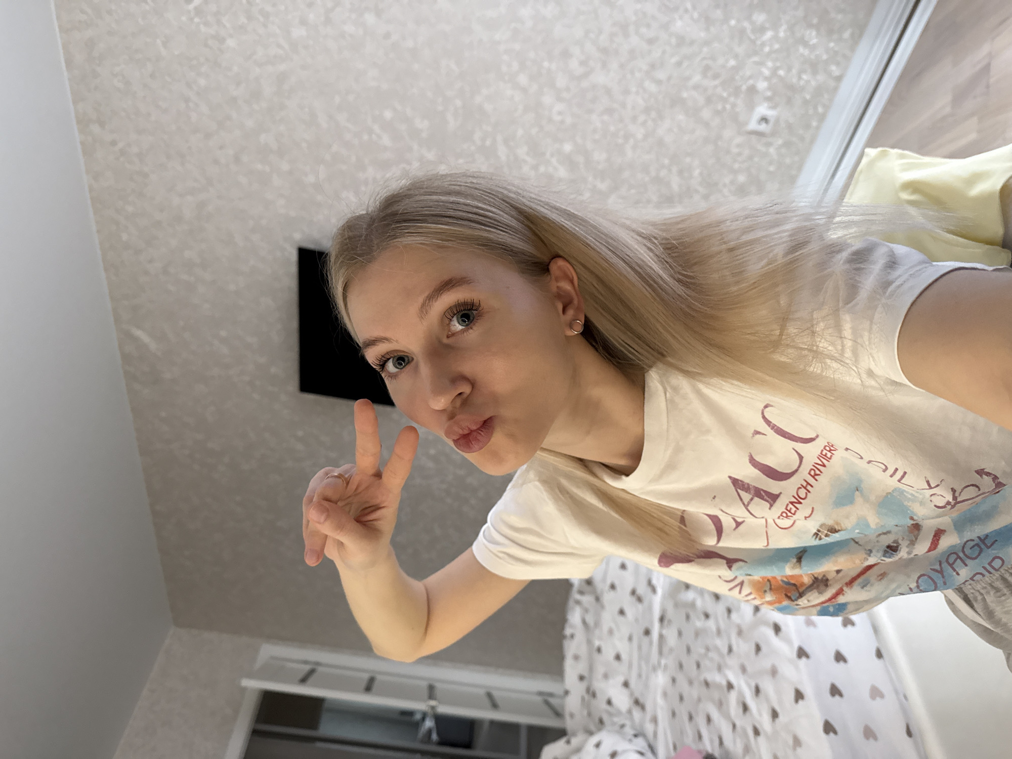 A young, blonde woman is taking a selfie in what appears to be her bedroom. She is holding up a peace sign with her left hand and puckering her lips. She is wearing a white t-shirt with a graphic print and her blonde hair is parted to the side. The room has a patterned wallpaper. There is a television mounted on the wall. The location details (Izhevsk, Russia) are not possible to ascertain from the image provided.