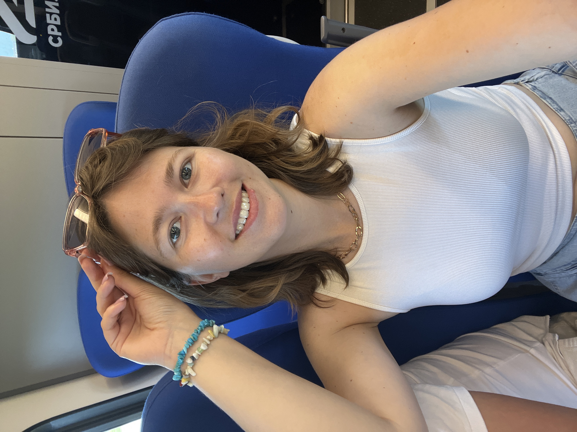 A young woman with light brown hair and blue eyes is posing for a selfie on a train in Zemun Urban Municipality, Serbia. She is wearing a white ribbed tank top, blue denim shorts, and has a gold chain necklace. She is holding pink-framed sunglasses in her right hand, and two colorful beaded bracelets are on her left wrist. She is sitting on a blue seat. The word "СРБИЈА" is visible in Cyrillic on the train.