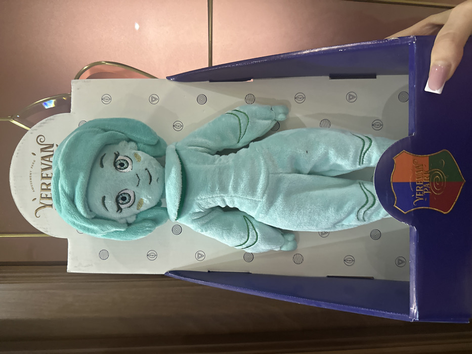 A light teal plush toy, approximately 30-40 cm in height, is displayed upright within its retail packaging. The plush depicts a stylized female figure with a large, rounded teal head covering, embroidered blue eyes with white pupils and black outlines, small eyebrows, two small golden-yellow embroidered dashes on each cheek, and a subtle stitched smile. The body is uniformly light teal, resembling a jumpsuit or robe, featuring a simple collar and green embroidered wavy lines on the upper arms and lower legs.
The packaging consists of a white cardboard back panel patterned with light grey geometric symbols. A rounded white header at the top reads "AMUSEMENT PARK" in smaller font, followed by "YEREVAN" in a larger, ornate gold script. The lower portion of the box, including the visible side panels, is dark blue/purple. Centered on the dark blue base is a multicolored shield logo (quartered in blue, orange, green, and red) with "Yerevan Park" inscribed in white.
A human hand, featuring a French manicure, is visible in the lower right, holding the dark blue base of the package. The background is blurred, showing a light pink surface with a subtle gold decorative element on the right, and a darker brown, possibly wooden, surface on the left. The item is identified as merchandise from Yerevan Park Amusement Park, located in Yerevan, Armenia.
