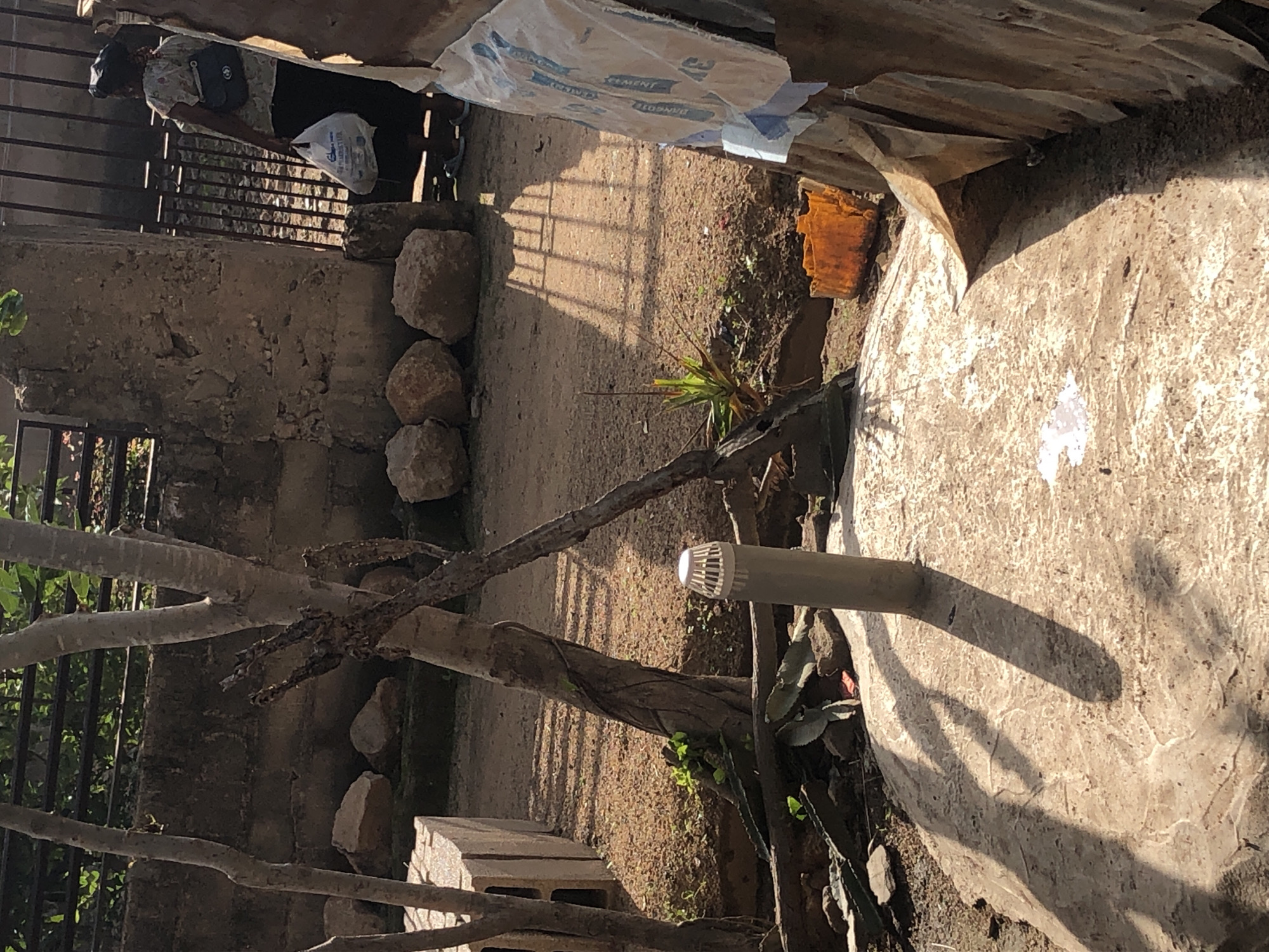 In a sunlit outdoor setting, likely a yard or alleyway, a person wearing a dark headscarf, light patterned blouse, and dark trousers stands near a metal gate. They carry a white plastic bag in their right hand and have a dark bag slung over their left shoulder. In the foreground, a round, textured concrete structure with a vertical pipe protruding from its center is visible. The ground is covered in dirt and scattered rocks. Shadows from a fence are cast across the ground. Partially obscured by the foreground structure is a corrugated metal siding. Behind the person and gate, a concrete wall and foliage are present.