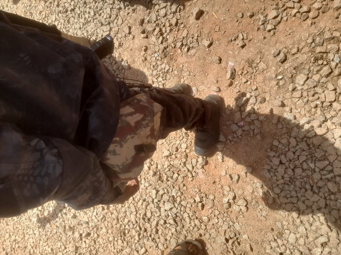 The image shows a lower-body view of an individual wearing a camouflage uniform, standing on a rocky, dirt surface. The individual's right foot is visible, and their left foot is partially obscured by the bottom of the image. The individual's right leg is wearing a dark boot. The individual is wearing a black jacket over a camouflage shirt. The individual's shadow is visible on the ground. The location of the image is a dusty road in Gusau, Nigeria (latitude: 12.156, longitude: 6.709). There are no other people visible in the image.