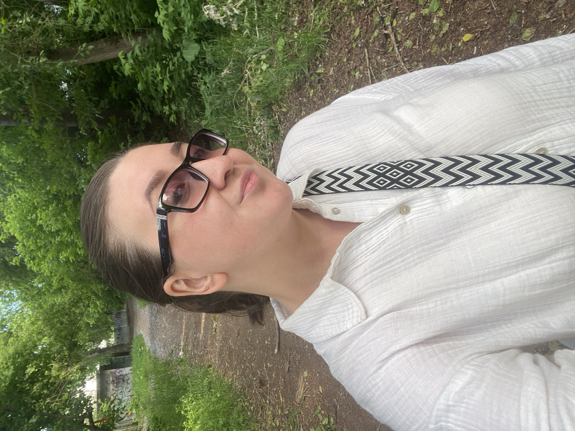 This vertical selfie captures a person outdoors in Chaikovsky, Russia. The subject is a person with dark hair tied back, wearing dark-framed glasses and a white, textured long-sleeved shirt. A black and white geometric patterned strap, featuring alternating chevrons and diamonds, is visible across their chest, partially tucked under the shirt collar. The person has light skin and a relaxed expression, looking slightly upwards towards the camera.
The setting is a natural, green environment, likely a park or a wooded area. The background is filled with lush green foliage from numerous trees and bushes. A dirt or gravel path extends into the distance behind the person. To the left side of the path in the background, there's a glimpse of a wall or structure covered with graffiti. The lighting suggests it is daytime, possibly under bright overcast skies or in soft sunlight, as there are no harsh shadows, indicating pleasant weather. No visible text is present in the image.