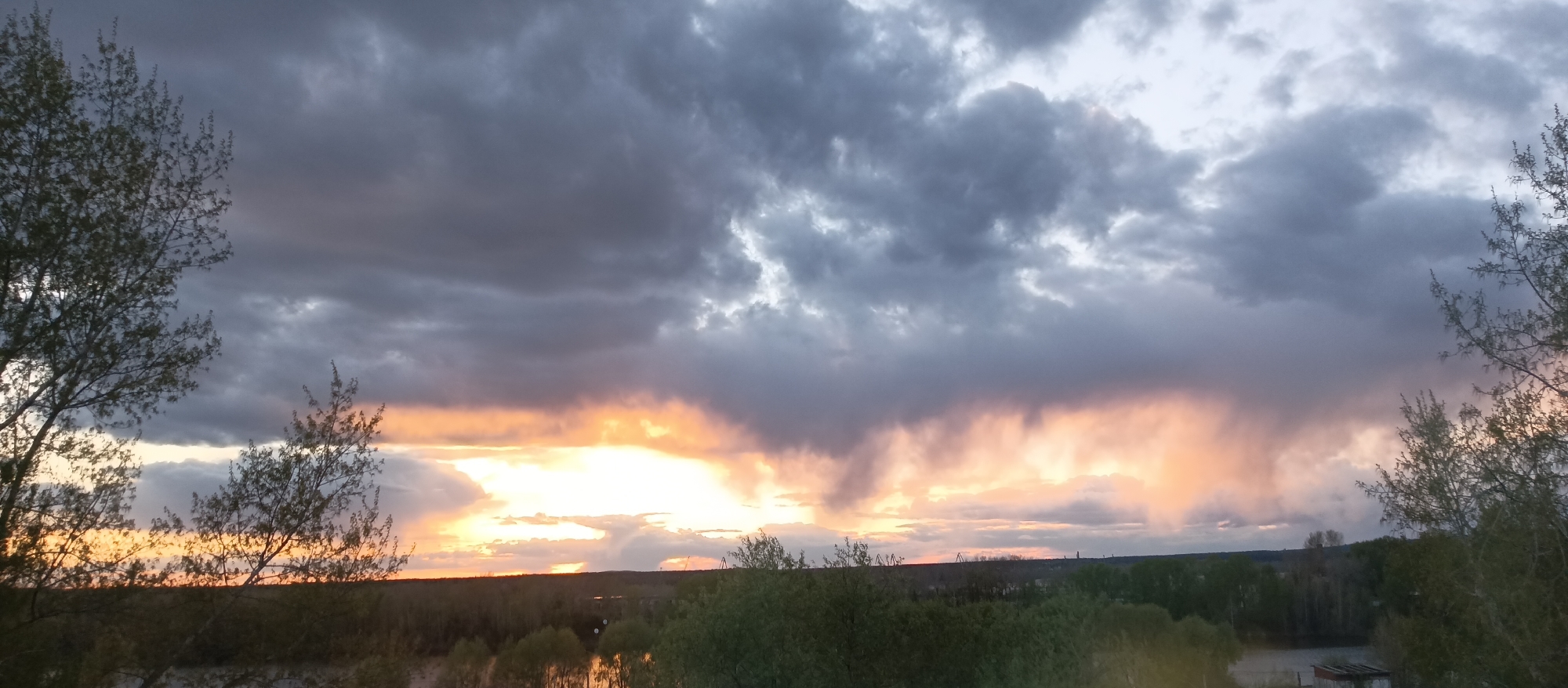 The image captures a dramatic sky with heavy, dark storm clouds dominating the upper portion. The sun is setting or rising, casting a bright, golden and orange glow that breaks through the clouds, especially in the central area. The light creates a striking contrast with the dark clouds above.
The foreground is filled with a landscape that includes trees and possibly a body of water, although details are somewhat obscured by the lighting and distance. There's a hint of a distant skyline with trees and structures visible along the horizon line.
The location appears to be an elevated viewpoint overlooking a natural landscape possibly within or on the outskirts of a city or town, based on the presence of structures in the distance.