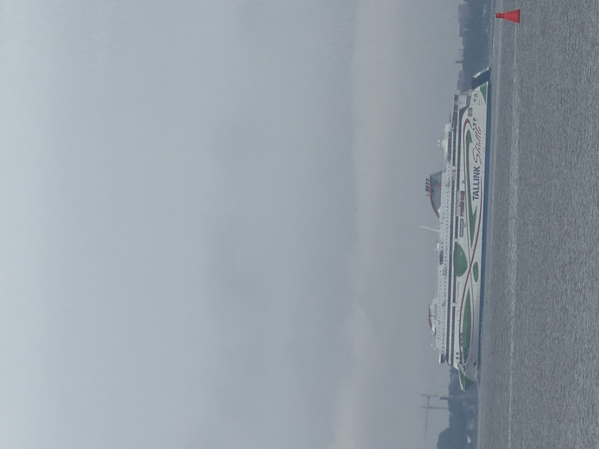 A large ferry, identified by the "TALLINK Shuttle" logo on its side, sails across a body of water. The ferry is predominantly white with green and red accents and designs. It is positioned horizontally in the frame, moving from left to right. In the background, a hazy cityscape is visible, with a bridge or structure on the left and more distant buildings on the right, suggesting a coastal or harbor area. A small, bright orange buoy floats in the water to the right of the ferry. The sky is overcast and grey, contributing to a muted overall color palette. The image appears to have been taken from another vessel or the shoreline.