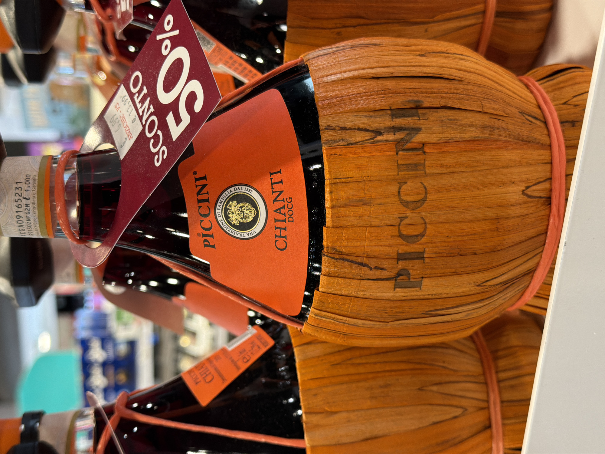 A close-up, eye-level, slightly upward shot focuses on three bottles of Piccini Chianti wine. The bottles are partially covered in a woven straw-like material and are tied with orange string. Each bottle has a prominent orange label with black text that reads "PICCINI" and "CHIANTI DOCG". A red sale tag is attached to the neck of the front bottle, advertising "SCONTO 50%" and listing the original price as €13,99 and the sale price as €6,99. The background is blurred, showing shelves stocked with other products, suggesting the bottles are on display in a retail environment. The lighting is bright, likely from overhead store lights. The overall impression is of a retail display in a store. The location is specified as Ferrara, Italy.