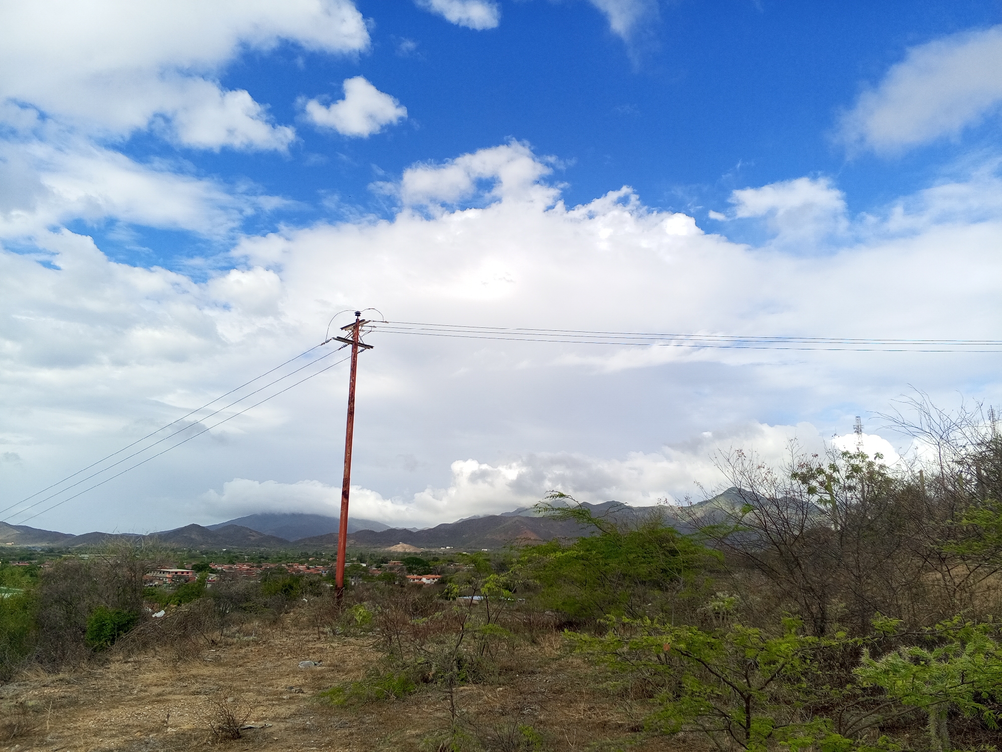 The image depicts a landscape near Juan Griego, Venezuela. The foreground is characterized by dry, scrubby vegetation on a brown, dirt-covered ground. A utility pole stands prominently in the midground, with power lines stretching across the scene. In the background, a town or settlement is visible, with buildings having reddish roofs. Beyond the town, hills and mountains are partially obscured by clouds, with some peaks shrouded in mist. The sky is partly cloudy, with patches of bright blue sky interspersed among white, fluffy clouds.