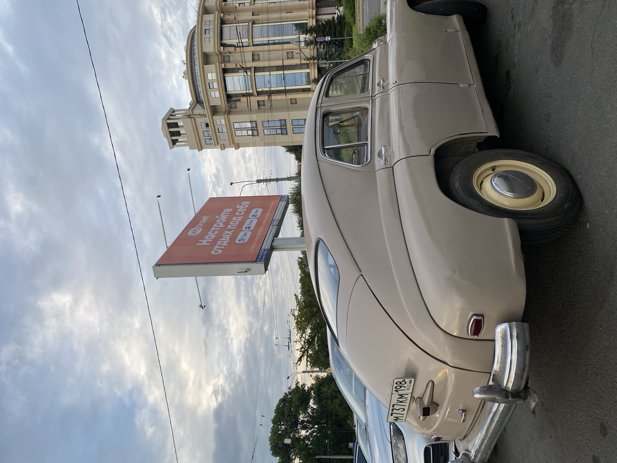 An aged, beige GAZ-21 Volga sedan is parked on an asphalt road in front of a modern, beige, circular apartment building. The car's rear is facing the camera, showing its chrome bumper and license plate reading "M737KM 198 RUS". A silver car is parked to the left of the Volga, partially visible. A large red billboard for "WB travel" stands behind the cars, with the Russian text "Настройте отдых под себя" (Arrange your vacation for yourself). The sky is overcast with scattered clouds, and trees are visible in the background. The scene appears to be an urban environment, likely in Saint Petersburg, Russia, given the presence of Russian license plates and Cyrillic text on the billboard.