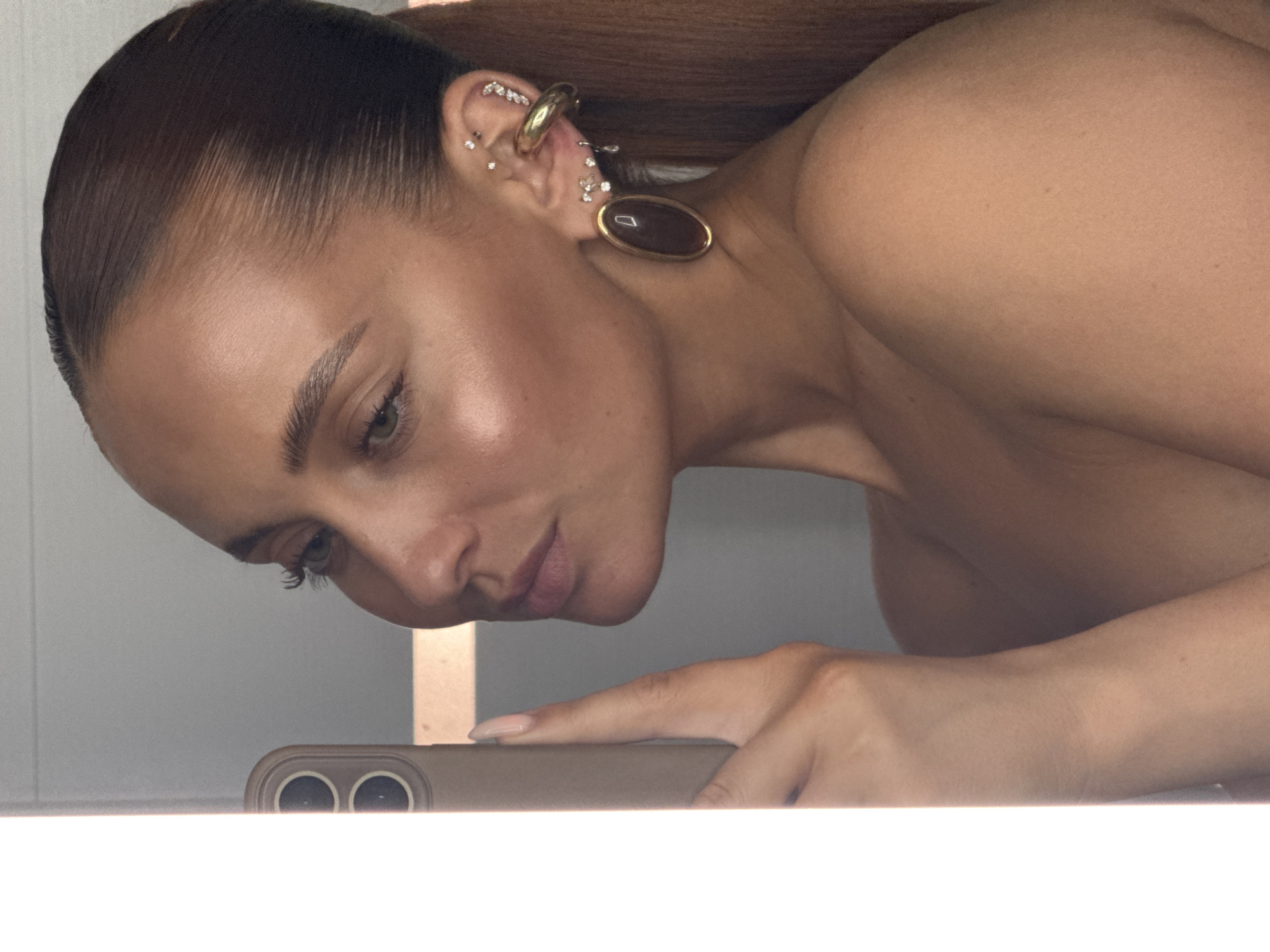 The scene is situated in a private interior space in the city of Chaikovsky, Russia. The image captures a female individual from the chest up, presented in a right-side profile as she gazes towards the left. She exhibits a deeply tanned complexion and sleek, dark brown hair meticulously pulled back into a high ponytail, visible over her right shoulder. Her facial features include green eyes, precisely defined eyebrows, and prominent cheekbones accentuated with a radiant highlight. Her lips appear full.
Her visible right ear is extensively adorned with multiple pieces of jewelry: a large, oval-shaped dangle earring featuring a dark, likely brown or black, stone encased in a gold-toned bezel. Above this, a thick, gold-toned ear cuff secures around the upper helix. Additional small stud piercings, each set with a clear, reflective stone, are arranged along her helix and lower earlobe, alongside a delicate, teardrop-shaped dangle earring.
The individual's right hand, displaying a long fingernail with nude-colored polish, holds a light-colored smartphone, identifiable by its dual camera lenses, positioned vertically to the left of her face. A intensely bright, vertical strip of white light borders the far left edge of the frame, originating from a significant light source or highly reflective surface immediately adjacent to the phone. The background consists of a plain, light grey wall, softly out of focus. The individual appears to be bare-shouldered.