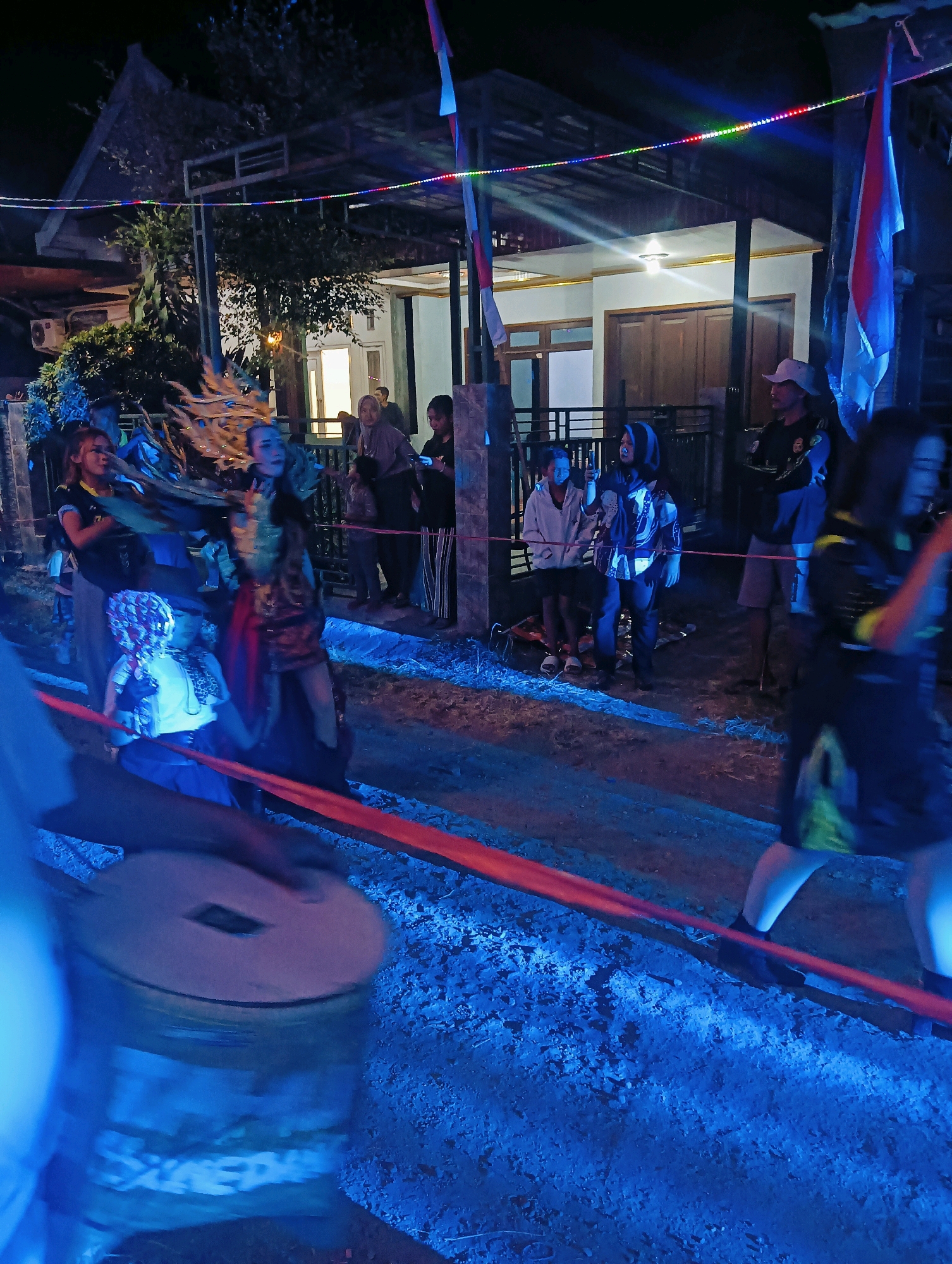 In Jatigedong, Indonesia, a nighttime cultural performance is underway. A group of individuals, some in elaborate costumes, participate in what appears to be a traditional dance or ritual. The scene is illuminated by decorative string lights and the lights from a nearby building, casting a blue glow over the unpaved ground. A red ribbon stretches across the foreground, suggesting a performance area or boundary. In the left of the frame, a person's arms are blurred as they play a drum. Spectators are visible in the background, observing the event.