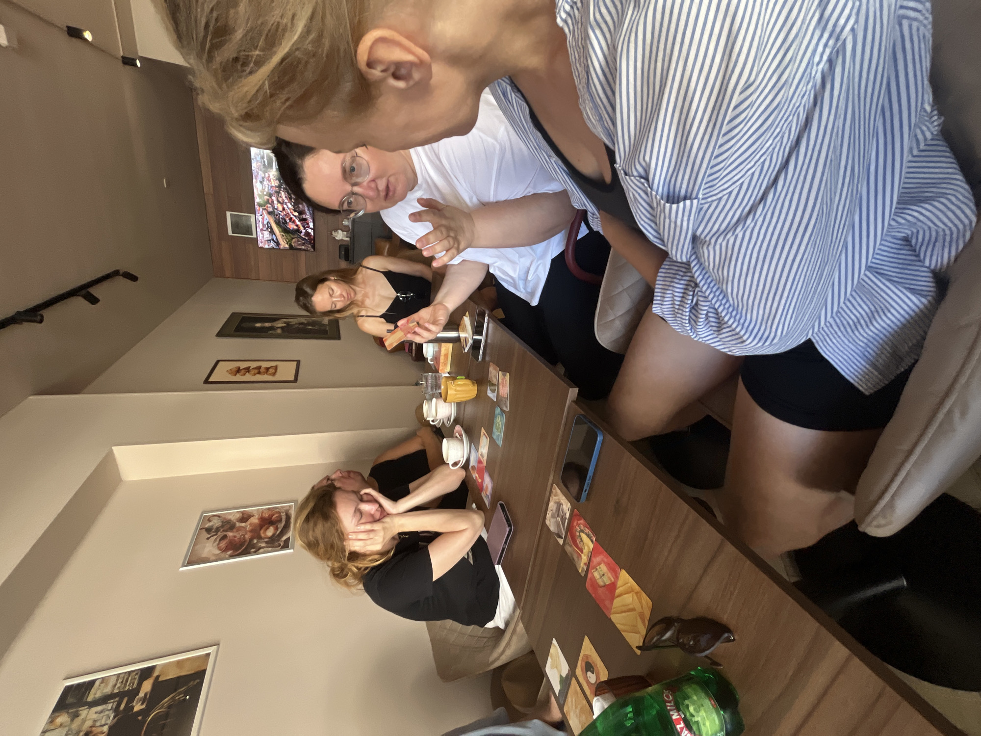 In Donji Pobori, Montenegro, at a cafe with beige walls and framed artwork, four women are seated at two adjacent wooden tables. The woman in the foreground, wearing a blue and white striped shirt, is partially visible, facing away from the camera and engaged in conversation with the woman next to her. This second woman, with dark hair and glasses, wears a white t-shirt and gestures with her hands as she speaks. To her left, a woman with blonde hair, wearing a black t-shirt, rests her head on her hands, appearing thoughtful or tired. Further down the table, a woman in a black tank top looks down at the table. The tables are set with coffee cups, saucers, a yellow mug, and several small cards arranged like a game. A green bottle of "Miloš" water and sunglasses are also visible on the table. A television screen is mounted on the wall in the background.