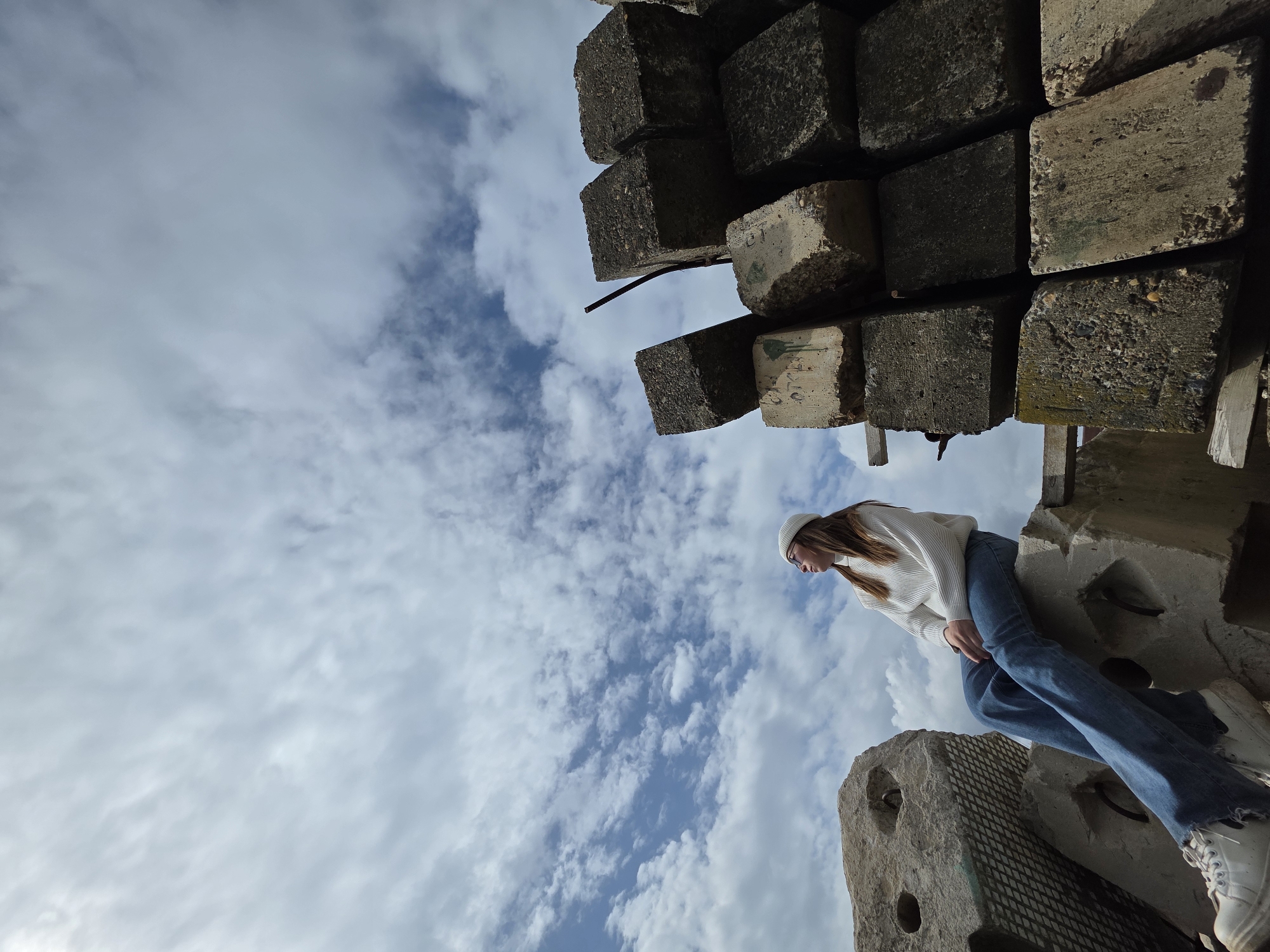 A low-angle shot depicts a woman perched on a stack of weathered concrete blocks. She wears a white sweater, blue jeans, a white beanie, and white sneakers. Above her, more concrete blocks tower against a sky filled with wispy clouds. The specific location within the city of Unknown, Unknown, is not discernible from the image.