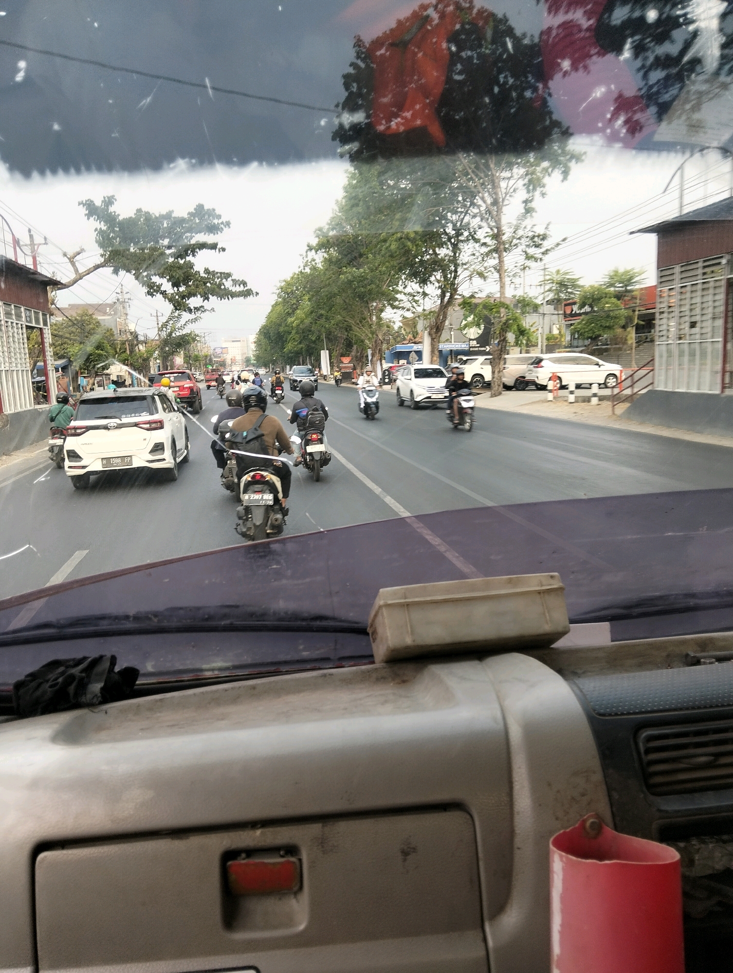 The image provides a forward-facing view from inside a vehicle, looking out through the windshield onto a busy city street in Semarang, Indonesia. The foreground shows the dashboard of the vehicle, with a beige rectangular object resting on top of it. The windshield itself is dirty, with reflections and streaks visible.
The middle ground of the image depicts a multi-lane road filled with traffic. Several motorcycles are the most prominent vehicles, with riders wearing helmets and casual clothing. Some cars are also present, including a white SUV in the left lane and other vehicles further down the road. Buildings line the right side of the street, with one structure featuring a prominent white grate-like facade. Trees with green foliage are present along the roadside and on the median.
The background shows more of the urban environment, with additional buildings and trees, and a cloudy sky. The overall impression is a typical street scene in a moderately trafficked Indonesian city. The presence of the Starbucks logo on a building suggests a commercial area within Semarang.