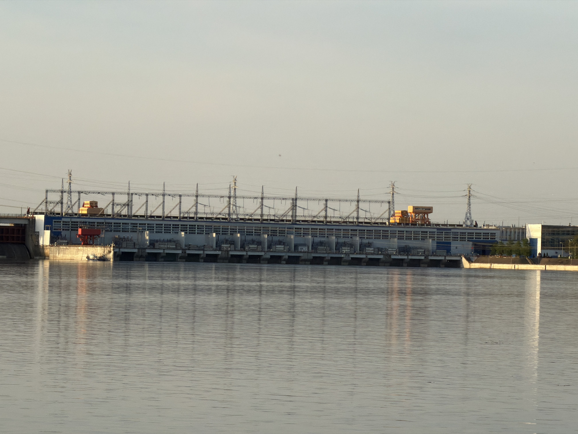The image depicts a hydroelectric power station on a wide river under a bright sky in Noviy, Russia. The station has a long, low building with a blue and white facade and a series of gates along the river. Above the building, there is a network of electrical transmission infrastructure, including pylons, transformers, and power lines. On either side of the main building, there are additional structures. The water reflects the station and sky, creating a mirroring effect.