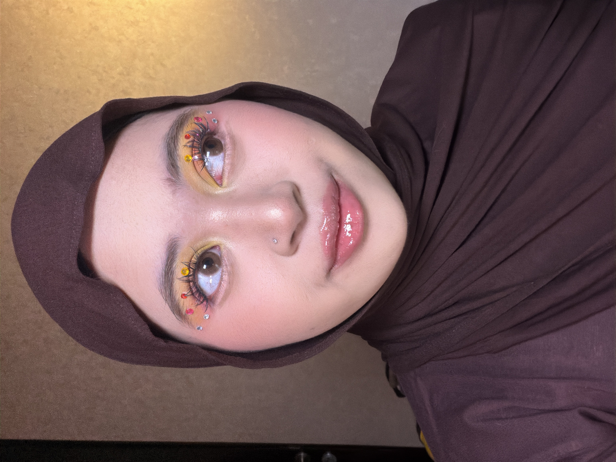 The media file shows a close-up, tilted portrait of a young woman wearing a brown hijab. She has striking eye makeup featuring yellow eyeshadow and a dramatic, multi-colored false eyelash application adorned with small rhinestones and colorful embellishments. Her lips are glossy and appear to be a light pink shade. A small silver stud is visible on her nose. The background is a textured gold surface, and the lighting suggests an indoor setting. The image does not contain any visible text.