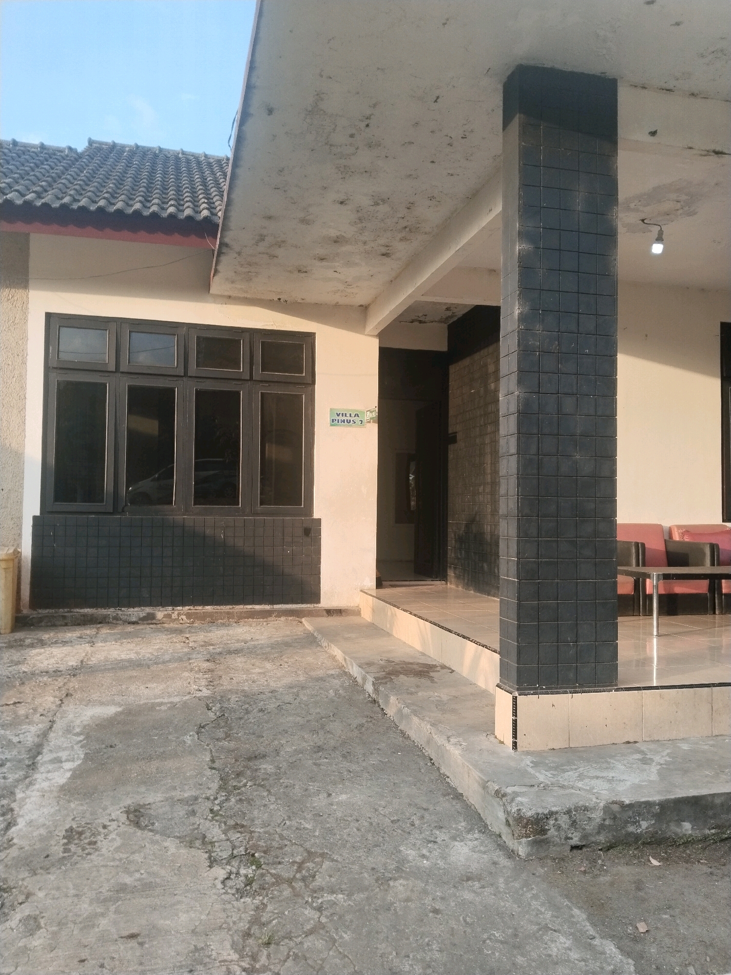 The image shows the exterior of a building, likely a villa, with a concrete pathway leading up to a covered porch. The building has a beige exterior wall and a dark-tiled base. A large dark-framed window with multiple panes is visible on the left side of the building. A sign that reads "VILLA PINUS 2" is mounted on the wall to the right of the window. The porch is supported by a large, dark-tiled rectangular pillar. Red and black chairs are visible on the porch, along with a small table. The ground in front of the building is paved with concrete, which appears cracked and weathered. The overall scene suggests an outdoor area of a residential or commercial property in a location that appears to be Guci, Indonesia, given the context provided. The sky is clear and light blue, suggesting daytime.