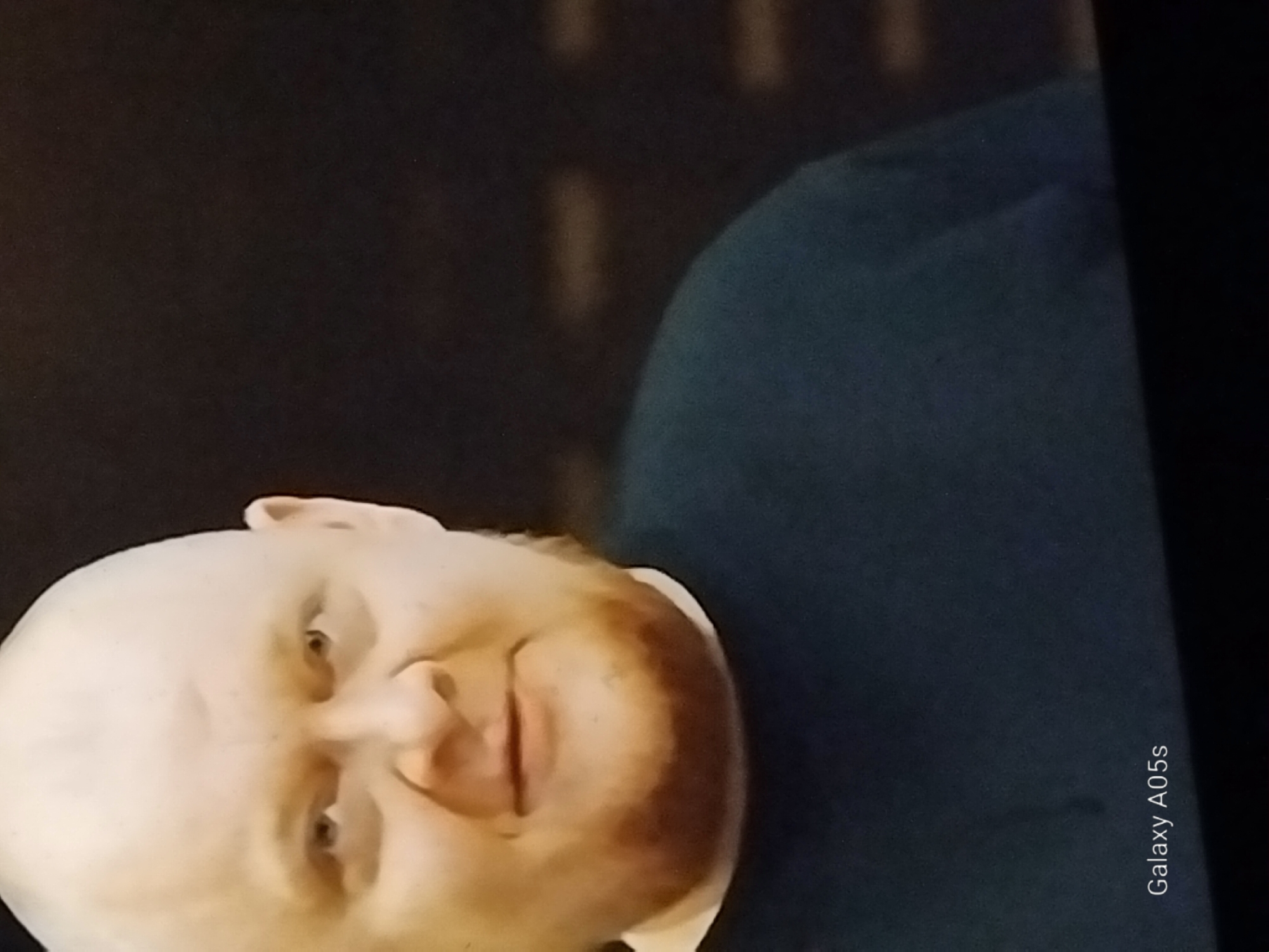 The image shows a close-up, head-and-shoulders shot of a bald, light-skinned man with a reddish-brown beard. He is wearing a dark blue crew-neck t-shirt. The background is dark and out of focus, with some lighter, rectangular shapes that could be lights or part of a textured wall. The lower left corner of the image has a watermark that reads "Galaxy A05s." There is no information in the image that would indicate the location in the city of Druzhba, Russia.