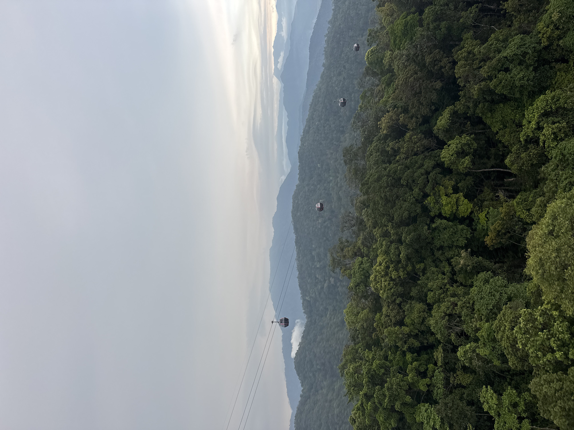 A slightly rotated, high-angle, long-shot captures a cable car system traveling over a lush, green mountain range. Four gondolas are visible at varying distances. The sky is overcast and white. The multi-layered mountain range in the background is lighter in color, receding into the distance. This is most likely a scene near Đà Nẵng, Vietnam.