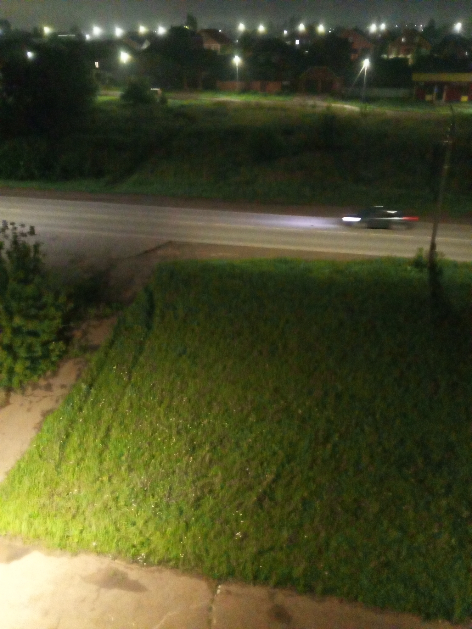 This is a nighttime, medium-angle, long-exposure photograph taken from a raised vantage point, looking down at a suburban street in Chistopol, Russia. The foreground is dominated by a gently sloping grassy area, illuminated by artificial light, with scattered small white flowers. The middle ground features a paved road, upon which a vehicle is visible as a blurred streak of light, indicating motion. The background shows a line of trees and houses, with streetlights casting a warm glow. The overall atmosphere is dark and quiet, characteristic of a residential area at night.
