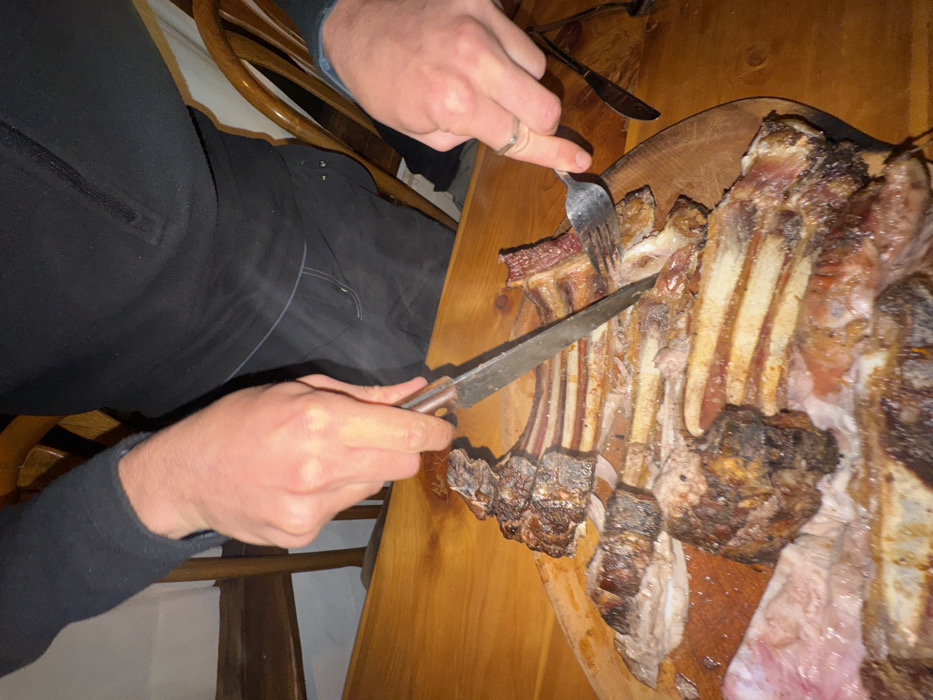 A person is using a fork and knife to cut into a rack of grilled ribs resting on a wooden surface. The person is wearing dark clothing. The ribs vary in color from light to dark brown and some red meat is visible. The wooden surface is light brown. A wooden chair is visible in the background.