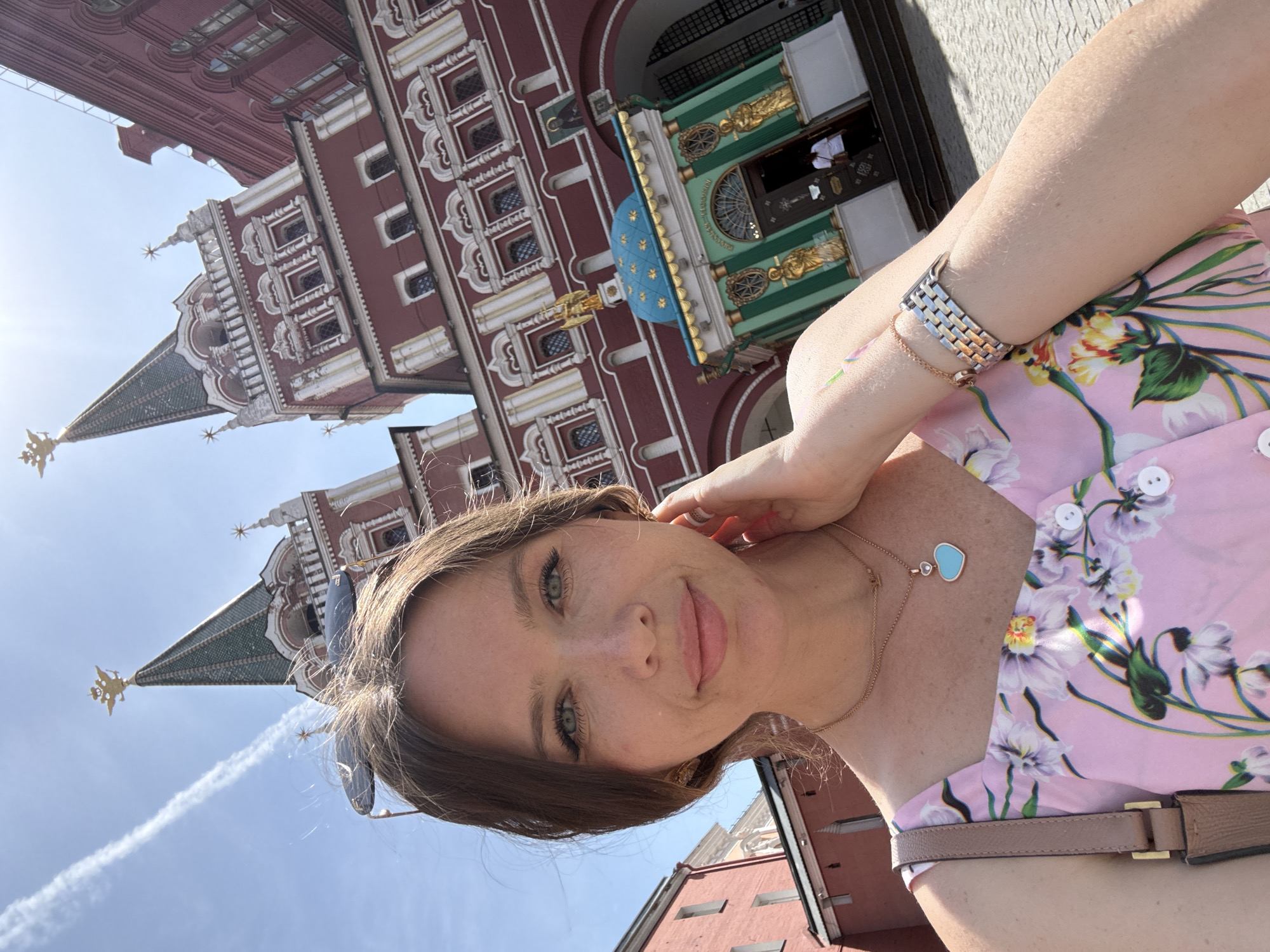 A young woman takes a selfie while standing in Moscow, Russia. The building in the background is part of Saint Basil's Cathedral and one of the towers is visible. The woman is wearing a pink floral dress, a watch, and a heart-shaped necklace. The sky is bright blue and there is a condensation trail behind an airplane.
