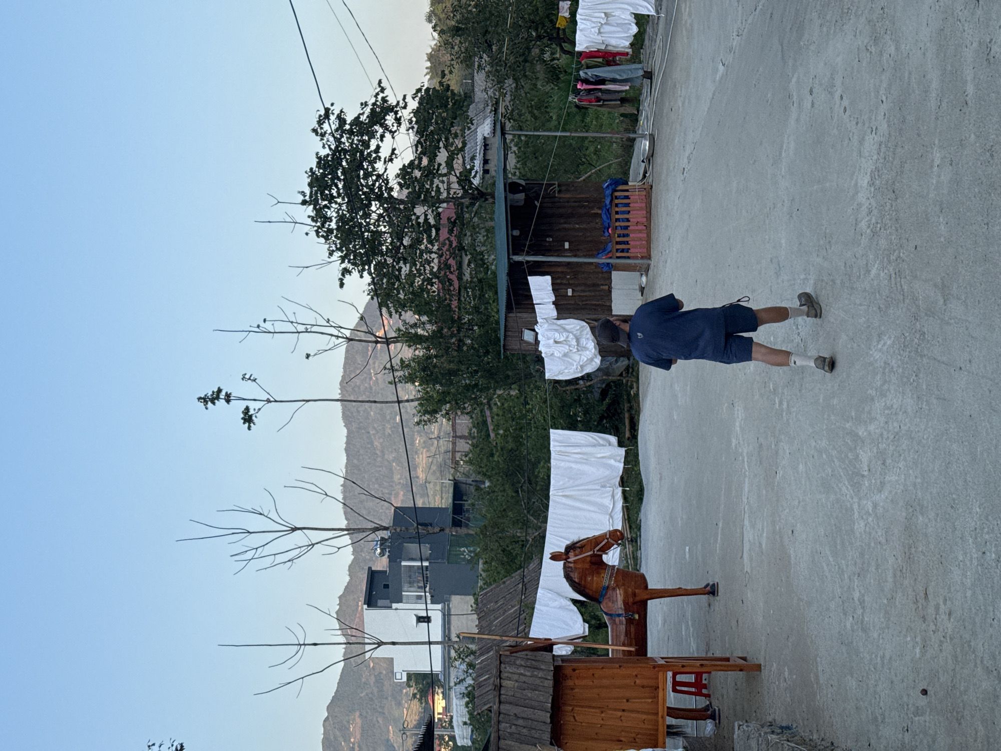 A rural scene in Bắc Hà, Vietnam, shows a concrete ground space with buildings and trees in the background. A wooden horse toy with a cart attached sits on the ground. Laundry hangs on clotheslines. A person is walking across the concrete. Mountains are visible in the distance under a clear sky. Power lines run overhead.