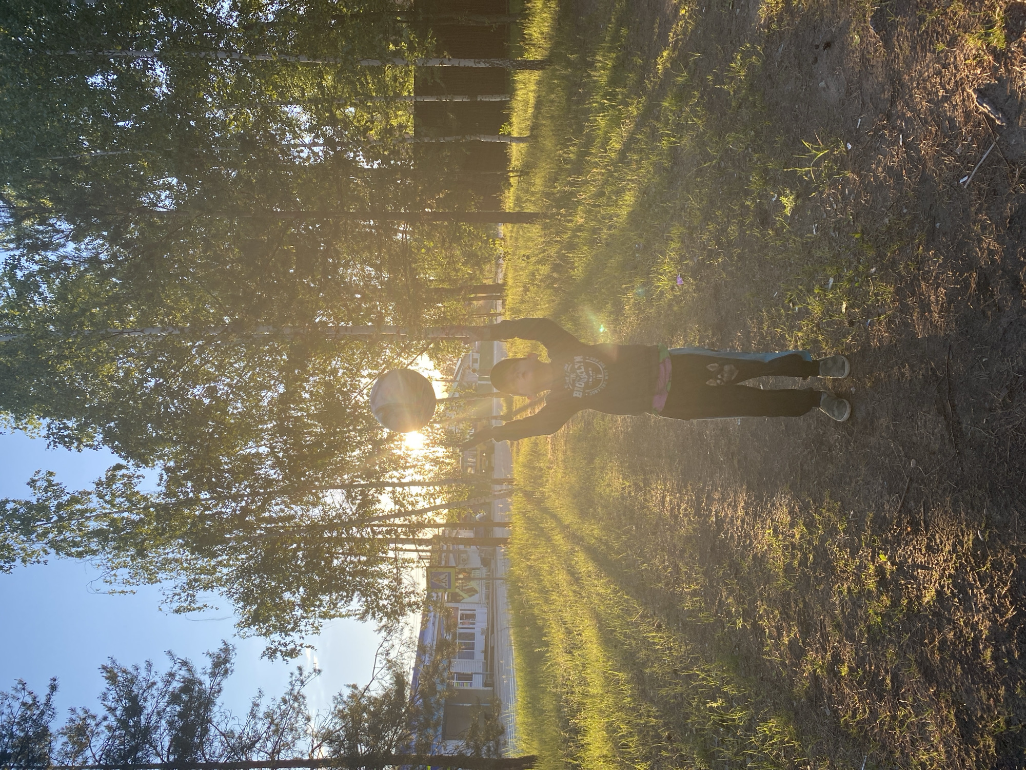 The image depicts an outdoor scene in Noviy, Russia, bathed in the warm, intense light of either early morning or late afternoon, creating a golden hour effect with prominent lens flare and long shadows.
The central subject is a young person, likely a child or teenager, seen from the waist up, actively engaged in playing with a multi-colored ball. They are wearing a dark long-sleeved top with the word "BROOKLYN" visible on the front, dark pants, and light-colored shoes. Their arms are extended upwards, indicating they are in the process of tossing or catching the ball, which is brightly illuminated by the sun directly behind them.
The setting is an open, natural area. Numerous trees with green foliage, appearing to be birches, line the background and mid-ground, casting long shadows across the ground. The ground itself is a mix of patchy green grass and bare earth. In the distance, behind the trees, several light-colored buildings and a yellow road sign are partially visible, suggesting a residential or semi-urban environment. The sky above is clear and blue. The scene conveys a sense of playful activity and bright natural light.