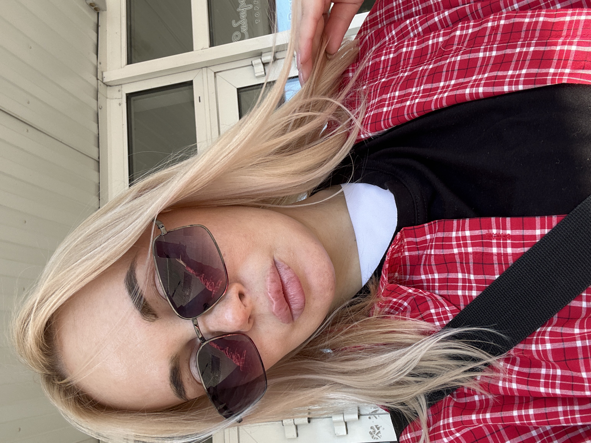 A woman is facing the camera, wearing sunglasses and a red and white plaid shirt over a black shirt with a white collar. She is holding her hair with her right hand. A black strap crosses her chest. Behind her is a white-framed door with rectangular panes. A building exterior is also visible. Based on the text visible on the door frame, the location is in Megion, Russia.