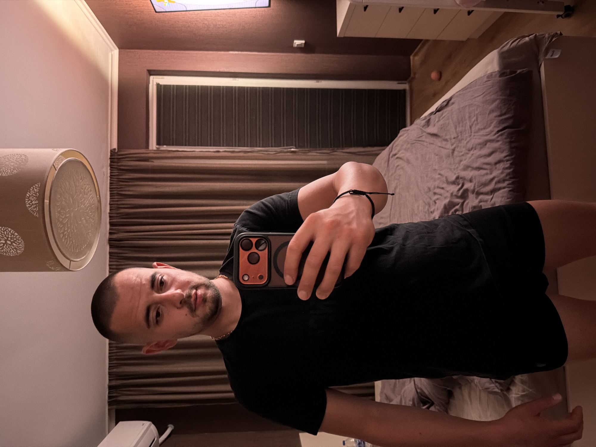 A man with a buzz cut and a short beard takes a selfie in a bedroom. He is wearing a black t-shirt and black shorts. He is holding a smartphone with an orange case. The room has brown curtains, a bed with a purple duvet, and a dresser with drawers. A basketball is on the floor near the bed. The lighting is dim, casting a pink hue on the walls. The scene appears to be in an apartment or hotel room. The presence of an air conditioner unit suggests an urban or suburban setting. No specific landmarks or details in the image indicate the city of Burgas, Bulgaria.