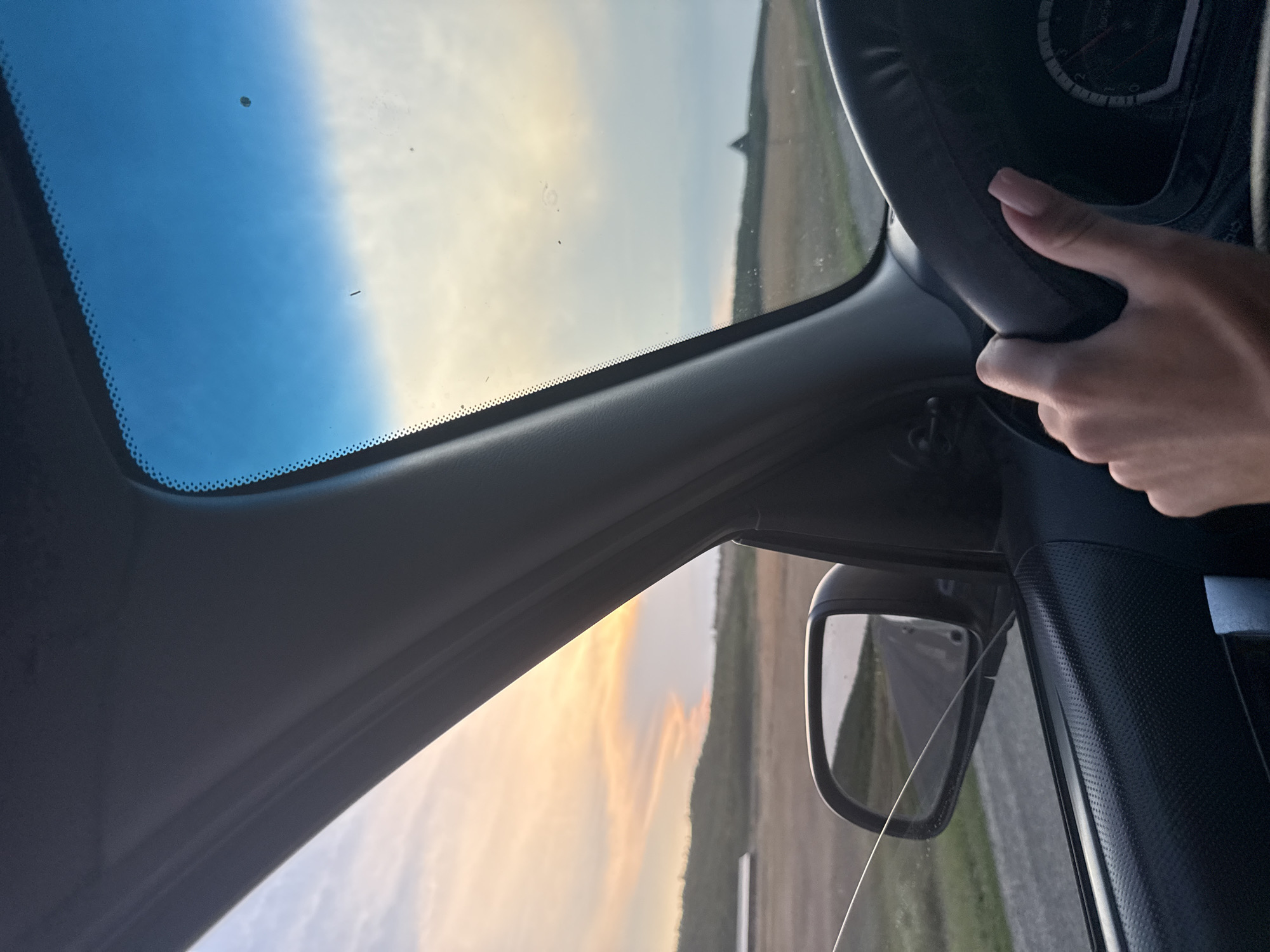 The image depicts a view from inside a car, with a hand on the steering wheel. The driver's perspective looks out through the windshield and side window towards a rural landscape at sunset. The sky displays hues of blue, yellow, and orange from the setting sun. Rolling hills with sparse vegetation are visible in the distance. A road stretches out, reflected in the side-view mirror. Part of the car's interior, including the dashboard and steering wheel, is in the foreground. The location is identified as Sadaclia, Moldova.