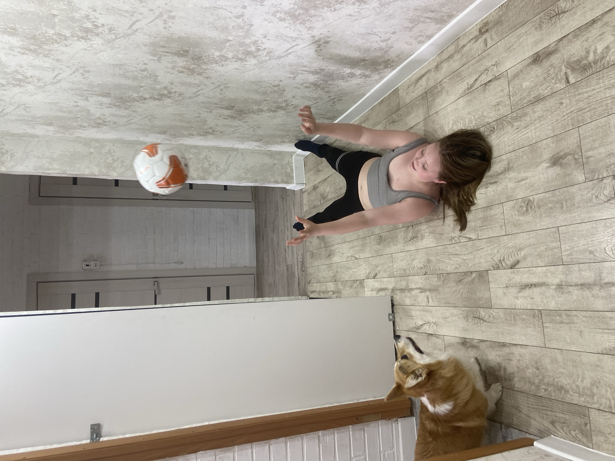 A young woman in a gray top and black leggings stands on a light brown wood-paneled floor. Her arms are raised, hands open, as if to catch a soccer ball hovering in the air. A corgi dog sits by a wall, looking upwards toward the soccer ball. The scene is indoors, and the location may be in the city of Chaikovsky, Russia.