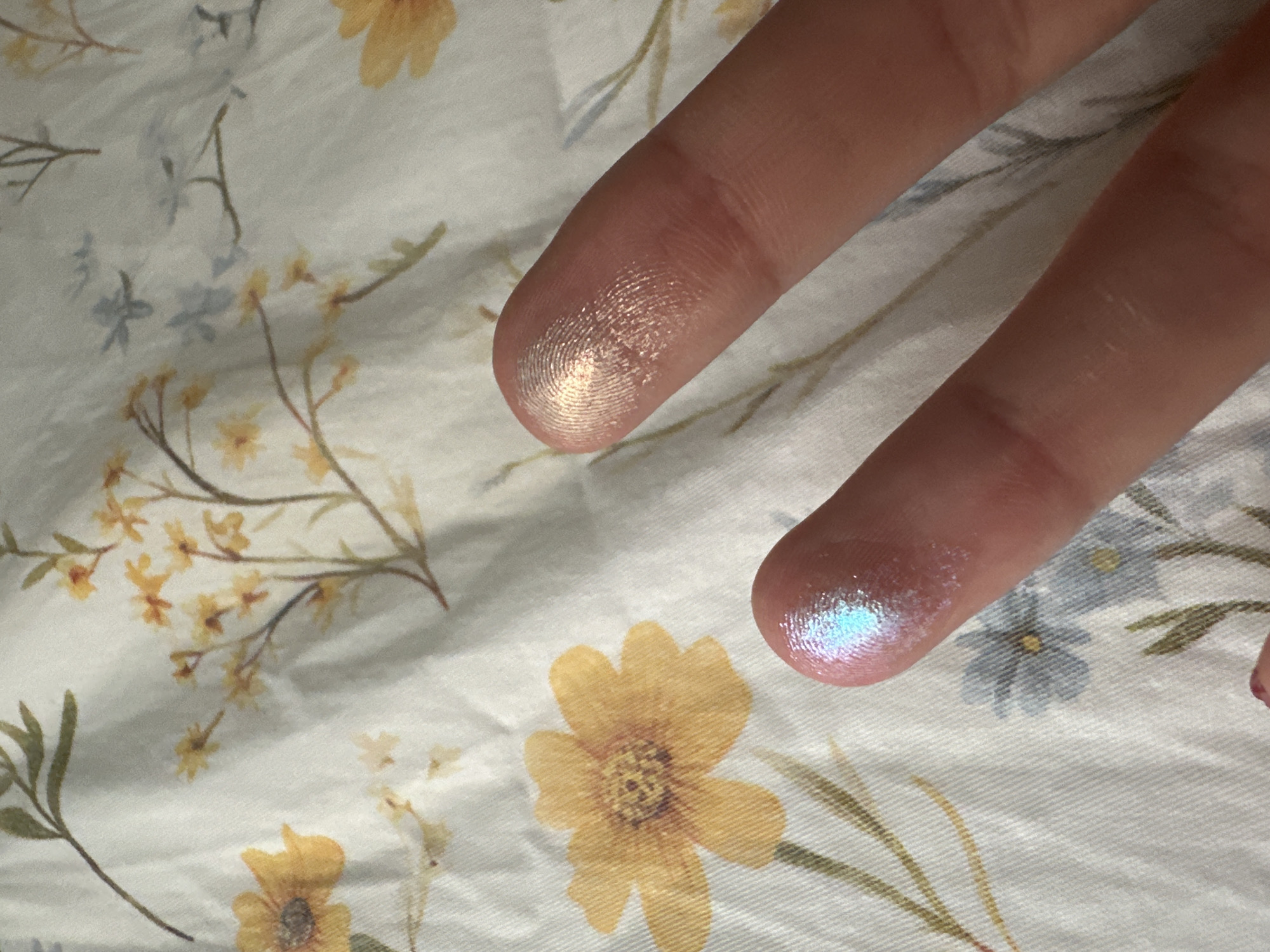 The image shows two fingers with shimmery cosmetic product on the fingertips. One fingertip has a gold-toned shimmer, while the other has a light blue shimmer. The background is a floral patterned fabric with yellow and blue flowers on a white base. The location is not discernible from the image content, therefore cannot be determined.