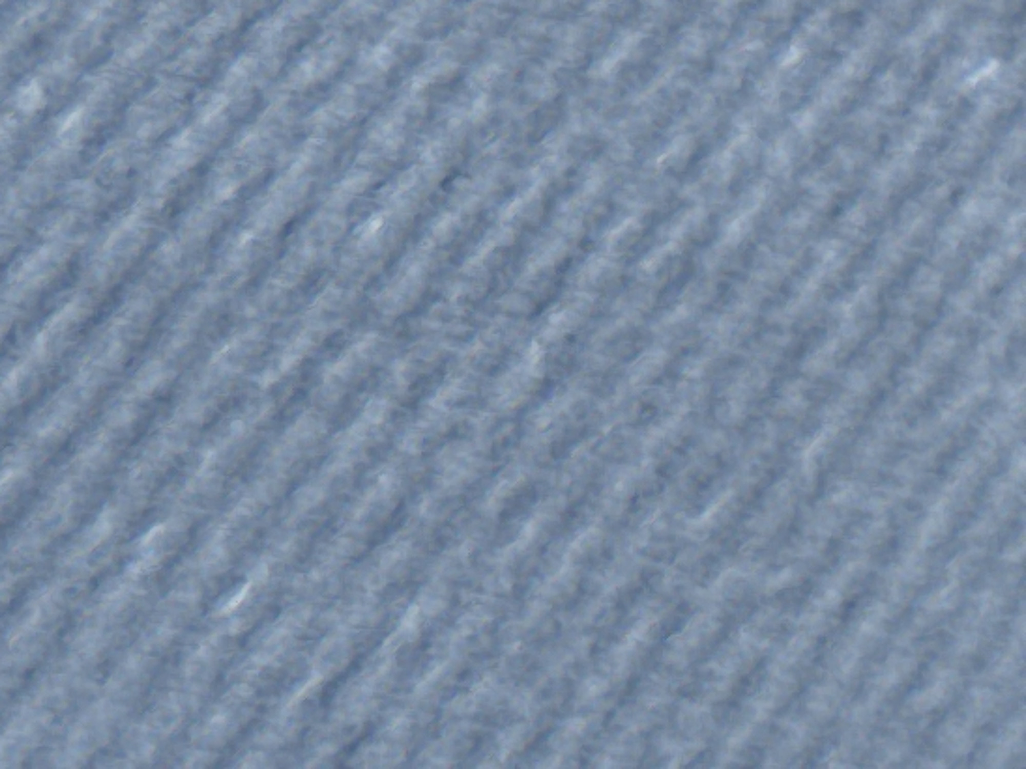 The image is a close-up, abstract view of a textured surface, likely fabric or a similar material. The texture consists of numerous parallel diagonal ridges, creating a ribbed or corded appearance. The color is a muted, light blue or grayish-blue. The lighting is diffused, with some subtle variations in shadow and highlight across the ridges, suggesting a soft, matte finish. There are no discernible objects, people, or specific environmental features visible that would allow for identification of a location in Varna, Bulgaria, or any other geographical context. The image focuses solely on the tactile and visual qualities of the textured material.