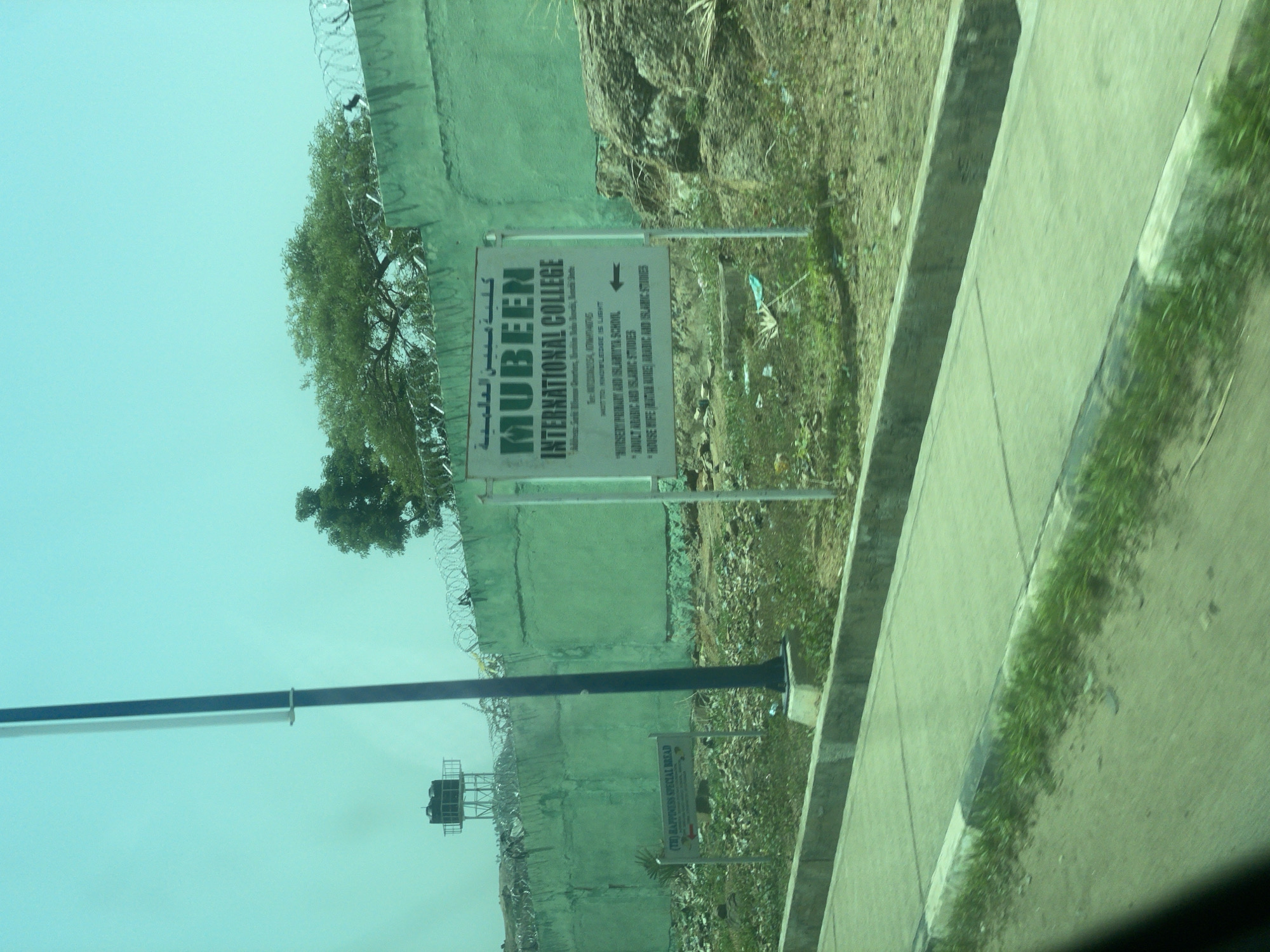 This image captures a slightly tilted view of a sign for "Mubeen International College" in Bauchi, Nigeria. The sign is white with green text and Arabic lettering at the top. Below the college name, it lists the school's motto as "Knowledge is Light" and provides details about "Nursery, Primary and Islamic School," "Adult Arabic and Islamic Studies," and "House Wife Juta Aure Maiic and Islamic Studies." An arrow points to the right, indicating a direction.
The sign is positioned alongside a green, textured wall, possibly a building or boundary, with barbed wire running along its top. To the left of the sign, a large tree with dense foliage is visible, its branches reaching towards the sky. Behind the wall, a rocky, overgrown area suggests a natural or undeveloped landscape.
In the background, a metal structure with water tanks is visible, possibly a water tower or part of a larger facility. The sky is a clear, pale blue, indicating daytime. The overall scene appears to be outdoors, by a road or pathway, with the green wall and surrounding vegetation suggesting a somewhat rural or semi-urban setting within Bauchi. The image is taken from a moving vehicle, as evidenced by the blurred foreground and the reflection on what appears to be a car window.