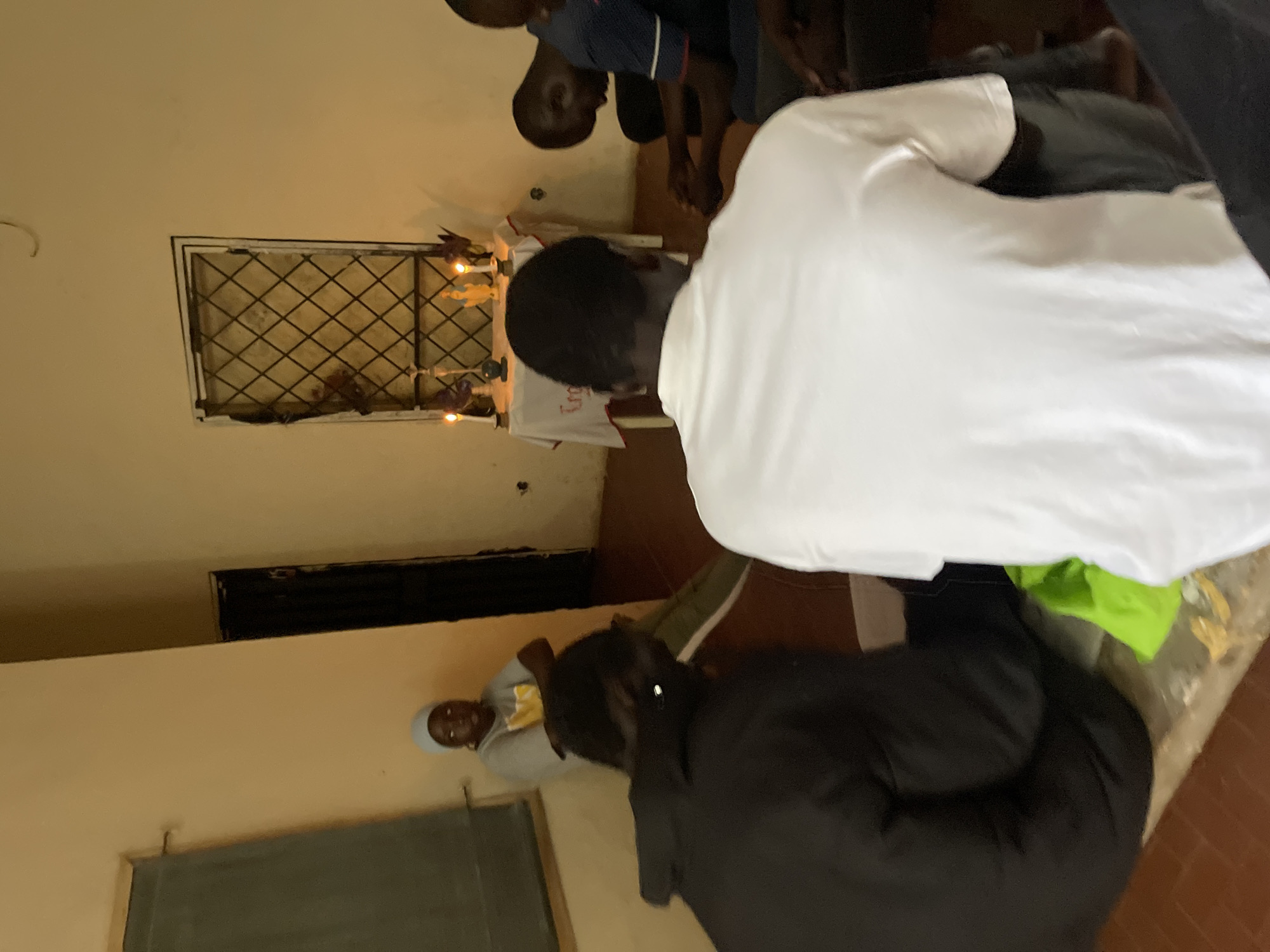 The image depicts a gathering of at least six individuals in a dimly lit room, possibly a community hall or a place of worship, in Rafin Par, Nigeria. The room has beige walls and a dark tiled floor. On the left side of the room, there's a dark-colored chalkboard or whiteboard mounted on the wall.
In the background, near a window with a diamond-shaped metal grate, a small table is covered with a white cloth. On the table, there are two lit candles, a small statue of a religious figure (likely the Virgin Mary), and some purple floral decorations. The presence of these items suggests a religious or spiritual gathering.
Several people are seen sitting on the floor, facing towards the table with the religious artifacts. One person, wearing a light blue head covering and a grey t-shirt with a yellow design, is sitting further back on the left, observing the scene. The majority of the visible individuals are men, with their heads bowed in what appears to be prayer or reflection. One person in the foreground is wearing a white t-shirt, and another is wearing a dark-colored jacket. A bright green bag is visible on the floor near the seated individuals. The overall atmosphere suggests a quiet and solemn occasion.
