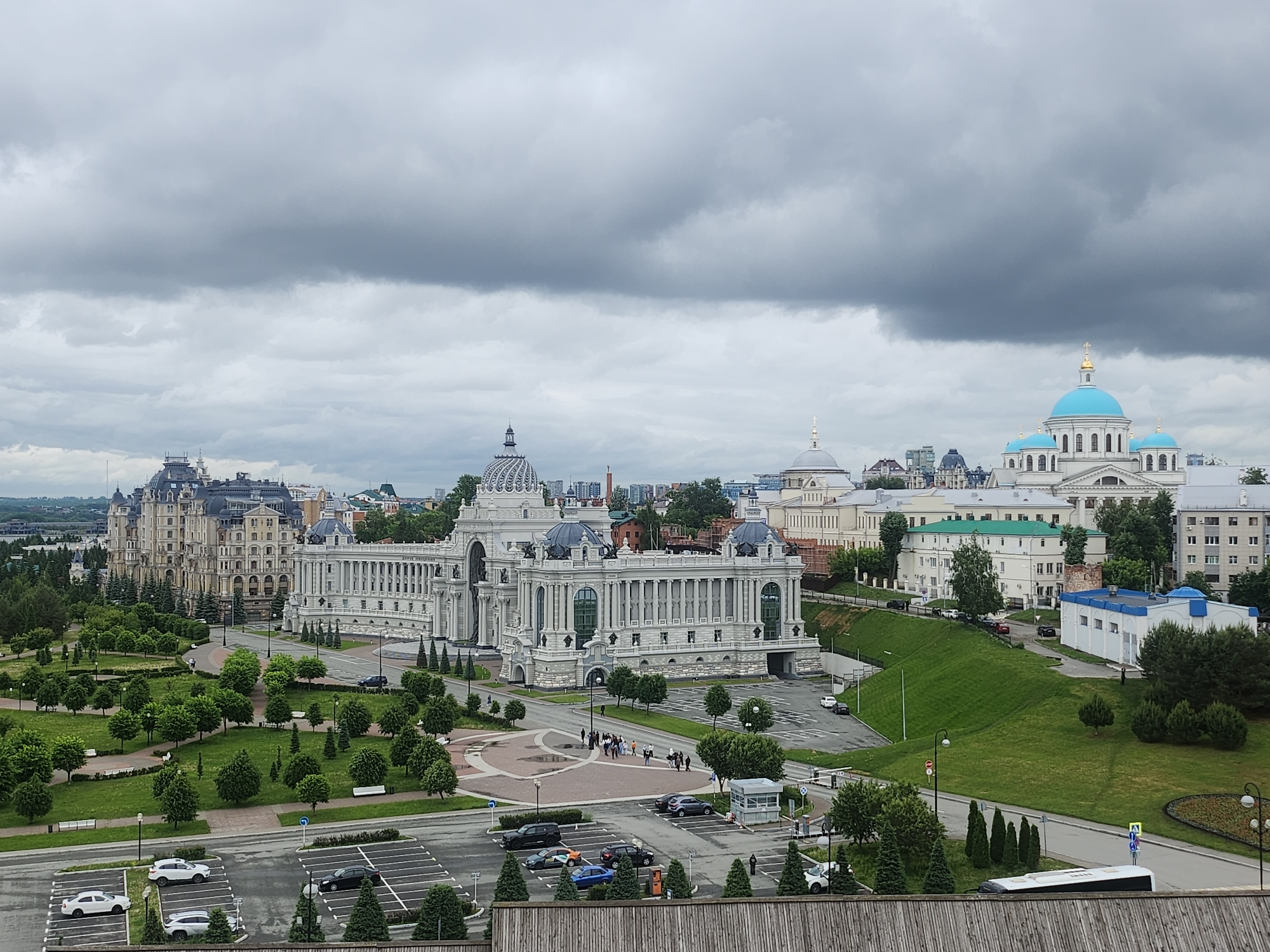 A wide-angle shot shows an urban landscape under a cloudy sky. The foreground contains a rooftop with a parking lot where various cars are parked. Adjacent to the parking lot is a green space with trees and benches. Behind this, an ornate, white building stands prominently, featuring multiple columns and dome-like structures. A group of people is visible walking near the entrance of the building. To the left, another grand building with a complex architectural design is present. In the background, additional buildings and domes are scattered across the city, with the blue domes of a cathedral standing out. The sky is covered with heavy, gray clouds, suggesting overcast weather.