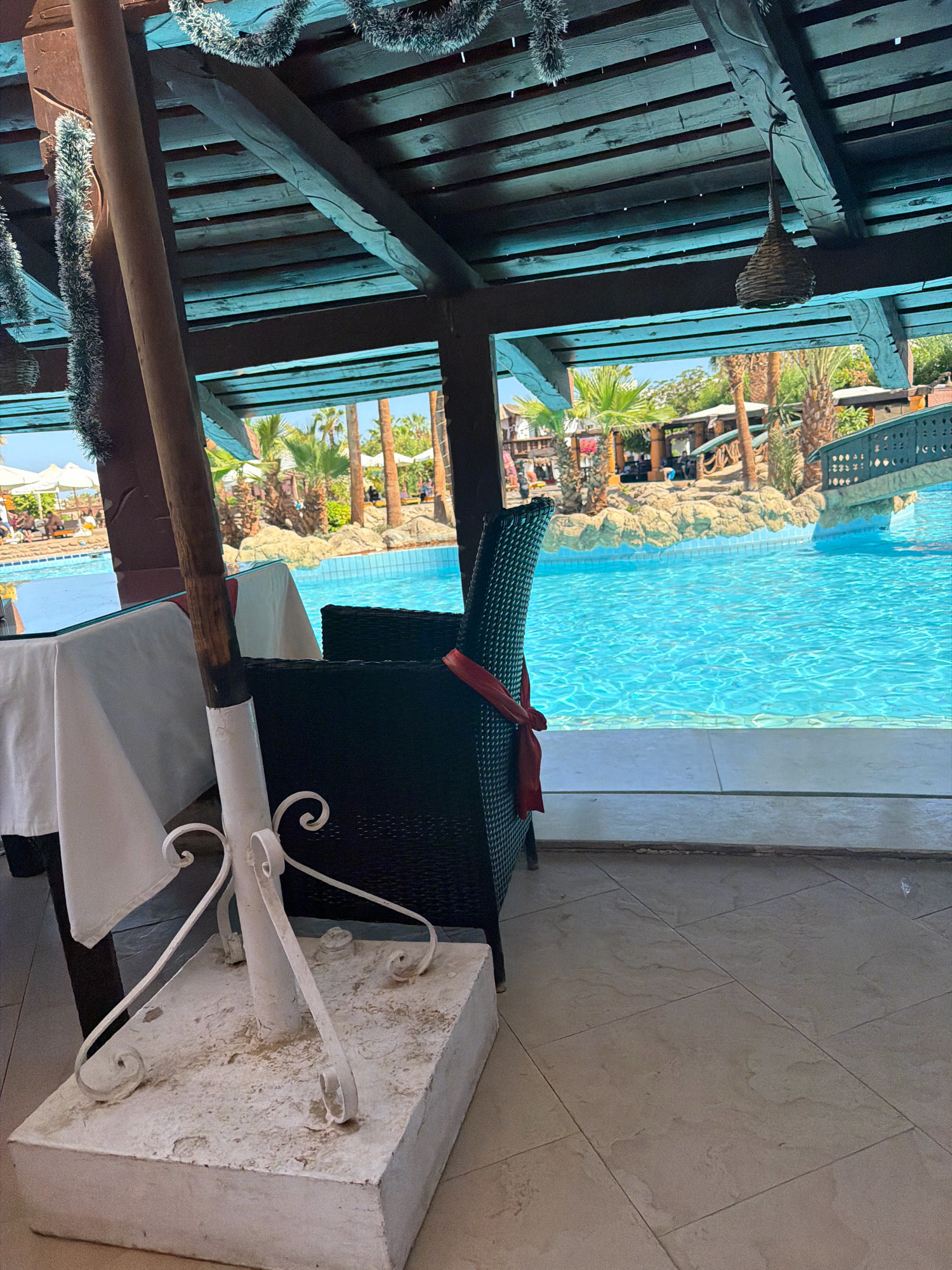 The image depicts an outdoor dining area adjacent to a swimming pool, likely located in Sharm El Sheikh, Egypt, given the tropical vegetation and architecture. In the foreground, a table draped with a white tablecloth sits to the left, with a white metal umbrella stand supporting a wooden pole in front of it. To the right of the table, a black wicker chair is visible, adorned with a red ribbon tied around its back. The floor is tiled with a light-colored, large-format tile. Beyond the seating area, a vibrant blue swimming pool dominates the scene. The pool is surrounded by decorative rocks and lush green palm trees, with a bridge arching over a section of it on the right side. In the background, more palm trees and some buildings with thatched roofs are discernible under a clear blue sky. Festive tinsel garlands are draped across the wooden beams of the overhead structure, suggesting a recent or upcoming celebration. The overall atmosphere is relaxed and resort-like.
