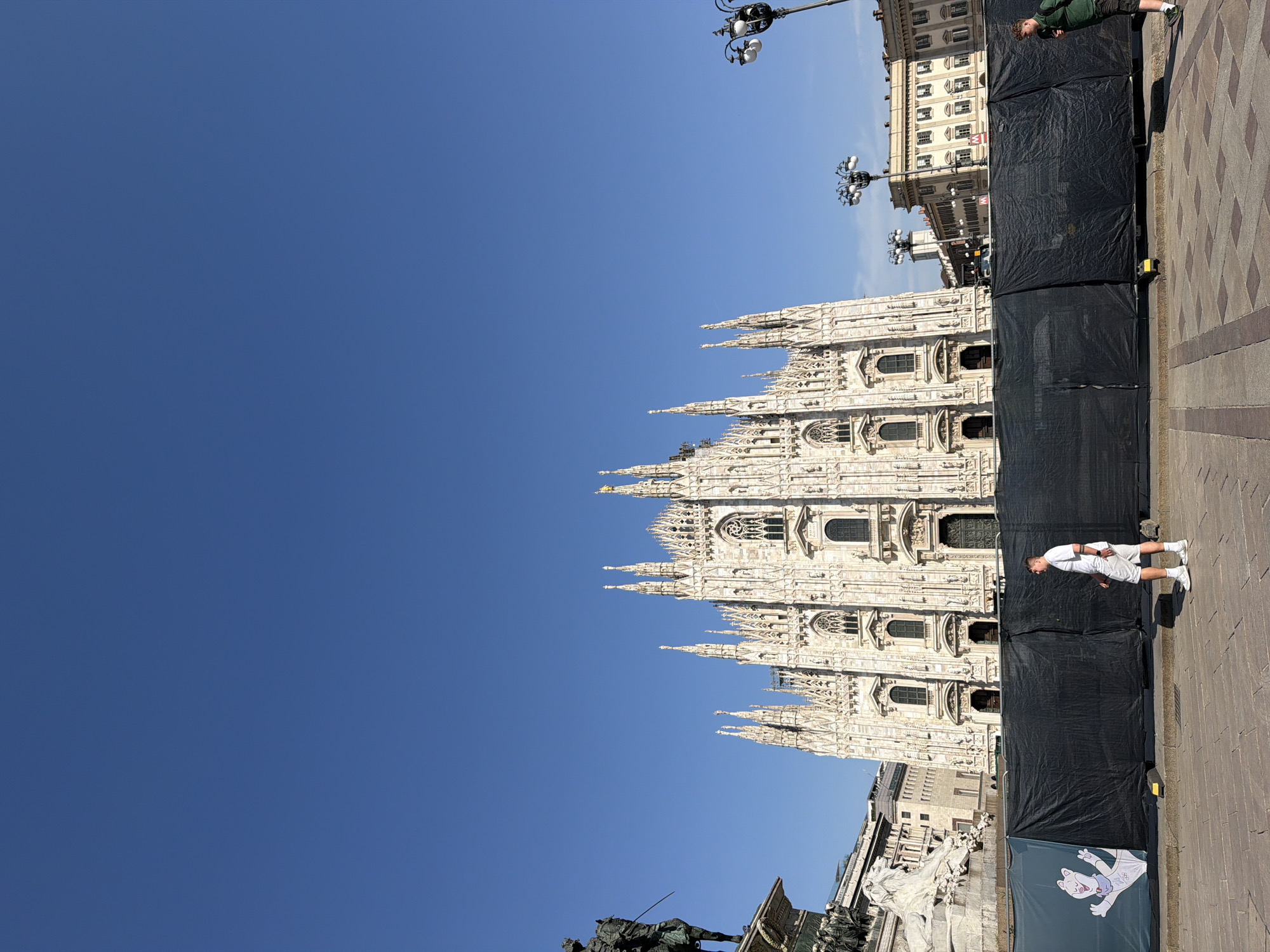 The image shows the Duomo di Milano, a large Gothic cathedral in Milan, Italy. The cathedral is made of white marble and features intricate spires and statues. The sky is clear blue. Part of the Duomo is obscured by black construction tarps. A person in a white outfit is walking near the tarps. A few buildings and street lights are visible behind the tarps. A statue with a man on horseback is in the lower left of the image.