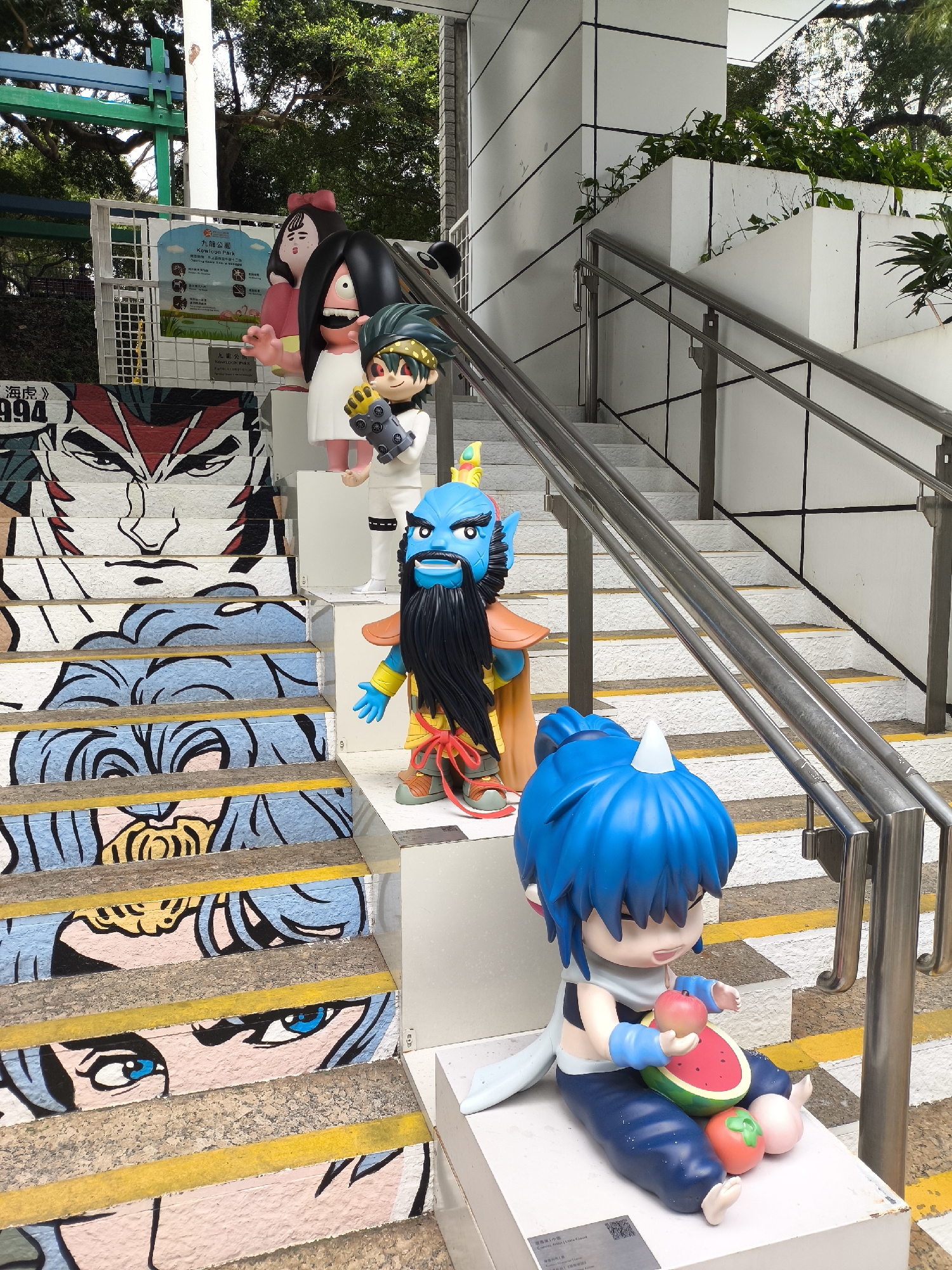 The image displays an outdoor public staircase art installation within Kowloon Park, Hong Kong, China.
The staircase is split visually; the left half features steps decorated with large, colorful pop art-style murals depicting manga/anime faces in shades of red, blue, black, white, and pink. Yellow anti-slip strips run across each step. A partially visible sign with "海虎》994" is affixed to the wall on the left. The right half of the staircase consists of white steps with yellow anti-slip strips and is bordered by a stainless steel handrail.
Five distinct, large-scale, stylized cartoon or comic book character figurines are positioned on white pedestals alongside the staircase, ascending from foreground to background on the right. From bottom to top, these include:
1. A seated chibi-style figure with bright blue hair, wearing a blue and white outfit, holding a red apple and a slice of watermelon, with a red tomato and a pink fruit near its feet.
2. A blue-skinned male figure with a long black beard, wearing a yellow and orange outfit with red accents, and a crown.
3. A male figure with spiky black and green hair, dressed in white with black accents, holding a yellow and grey mechanical device.
4. A pale figure with black hair, a white dress, and a wide-open mouth displaying teeth, with a large yellow circular left eye.
5. A large female figure with black hair, a red bow, and a pink dress, with outstretched arms.
In the upper left background, dense green foliage and trees are visible, along with a blue and green structural element. A prominent white sign with a blue border, stating "九龍公園 KOWLOON PARK" in Chinese and English, displays pictorial park rules. The upper right background shows a modern white building facade with dark vertical and horizontal lines, and planted greenery.