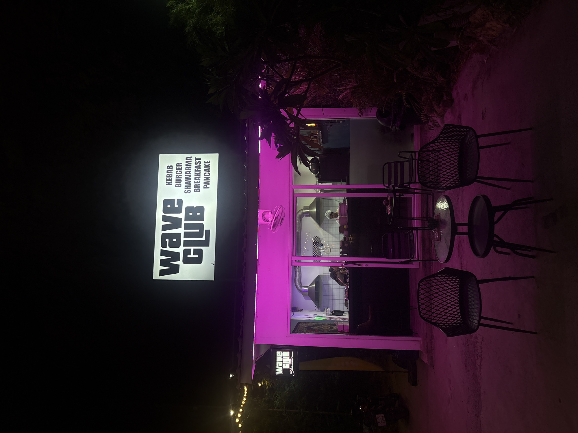 At night in Baan Bang Rak, Thailand, a "Wave Club" restaurant is illuminated with purple lighting. The restaurant's menu lists kebab, burger, shawarma, breakfast, and pancake options. Outside, there is seating with black metal chairs and a small round table. Inside the restaurant, one person is visible in the kitchen area, possibly preparing food. A small string of lights is visible further down the street, and a motorcycle is parked nearby. The environment is dark with some foliage visible.