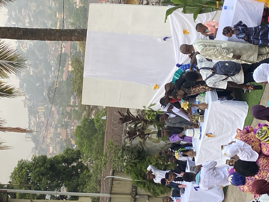 An outdoor social gathering is depicted on an elevated terrace or balcony, overlooking a sprawling, densely populated hilly cityscape, characteristic of Freetown, Sierra Leone. The scene is under soft, diffused lighting, suggesting late afternoon or an overcast day.
Numerous individuals, mostly adults with a few children, are present, seated and standing around white-covered rectangular tables. The attire is diverse, including traditional African garments such as men's long kaftans (some in light tones, others in olive green) and women's vibrant patterned dresses, mixed with more casual clothing. Many men are wearing white prayer caps or kufis.
The tables are draped with white tablecloths and hold bottled water, plastic cups, and decorative elements. A small white two-tier item with blue accents, possibly a cake or centerpiece, is visible, along with arrangements of yellow artificial flowers. White fabric panels serve as a backdrop or overhead covering, decorated with small yellow and blue ribbons or details. Lush tropical vegetation, including a large palm tree trunk and fronds on the left, and banana leaves behind one table, contributes to the outdoor setting.
Attendees are engaged in various activities, including socializing and interacting with items on the tables; some individuals are looking at their mobile phones. The overall atmosphere appears relaxed and communal, indicative of a special occasion or social event. The background reveals numerous buildings scattered across the hazy hillsides under a pale sky. No legible text is visible on any objects or clothing.