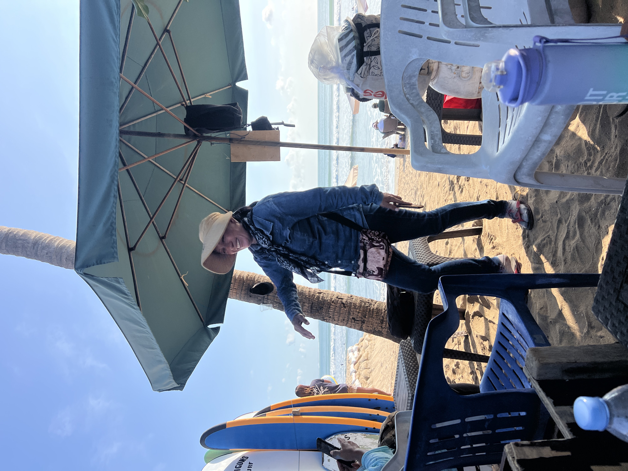 A woman wearing a straw hat and denim jacket stands on a sandy beach in Kuta, Indonesia, under a large green umbrella. She is gesturing with her right hand and smiling. Behind her, surfboards are lined up, and the ocean with gentle waves is visible. In the foreground, several plastic chairs and a table are set up on the sand. A palm tree trunk is to the left of the woman. The sky is blue with scattered clouds.