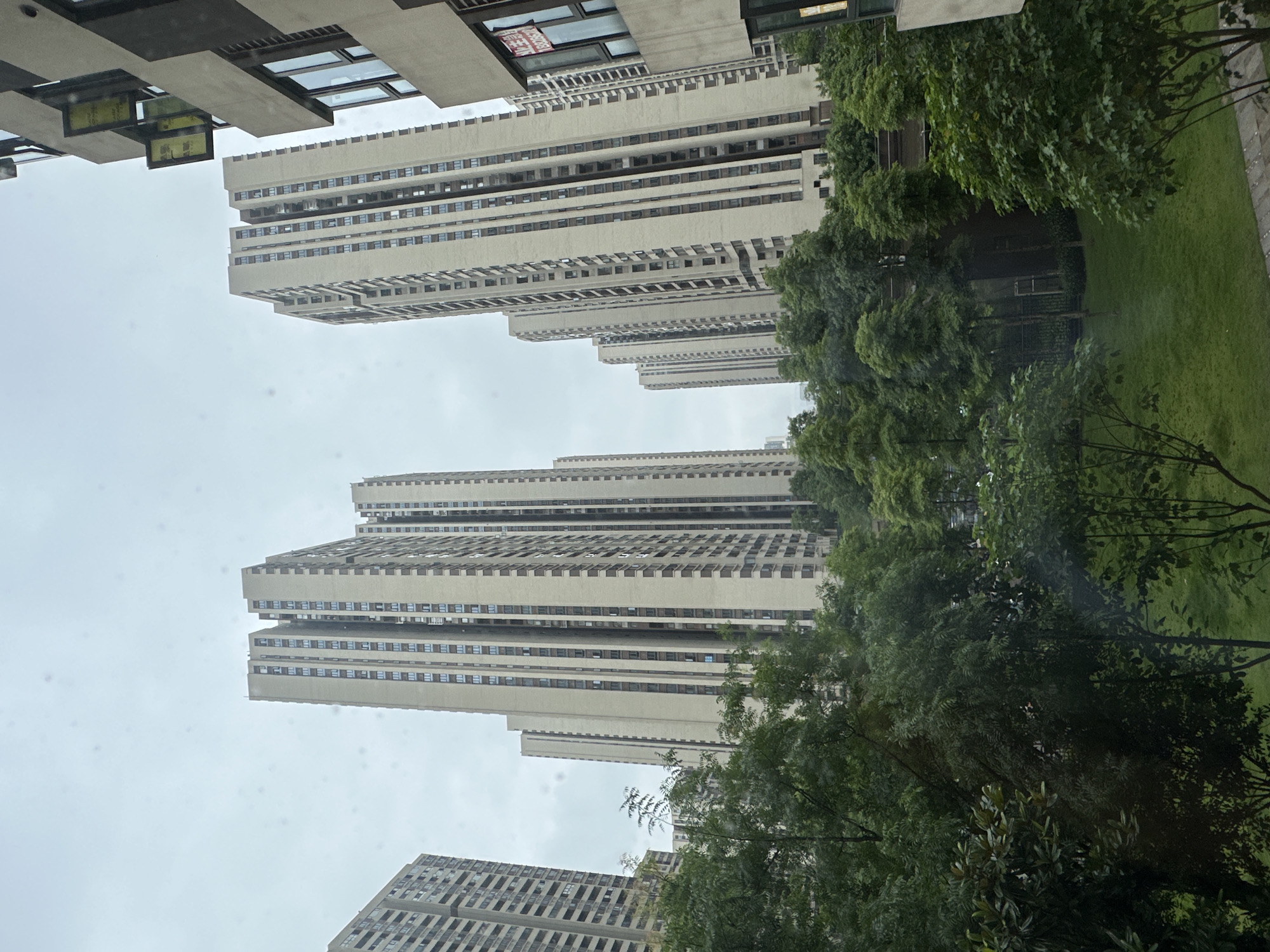 This image captures a residential urban landscape in the Chongqing High-tech Zone, China, under an overcast sky. The scene is dominated by multiple tall, slender high-rise apartment buildings, characterized by light beige or cream-colored facades with darker vertical elements and window frames. These structures exhibit a repetitive architectural design, receding into the background, signifying a densely developed residential complex.
In the midground and foreground, a lush green environment is prominent. Various deciduous trees, ranging from dense canopies to smaller saplings, fill the lower half of the frame with dark green foliage. A well-maintained patch of vibrant green grass, indicative of a lawn or park area, is visible in the lower center-right. A paved edge or pathway is partially discernible at the bottom right, bordering the grassy area.
On the far right, a portion of a closer building, sharing a similar light-colored facade, is visible. One of its windows displays a red sign with white Chinese characters that read "租售" (rent/sale) and a partial phone number, "180830...". Slightly above this, two yellow rectangular banners or awnings project from the building's exterior. The sky is a uniform light grey, indicating heavy cloud cover and resulting in soft, diffused lighting across the entire scene. No individuals, animals, or specific actions are observable within the frame.
